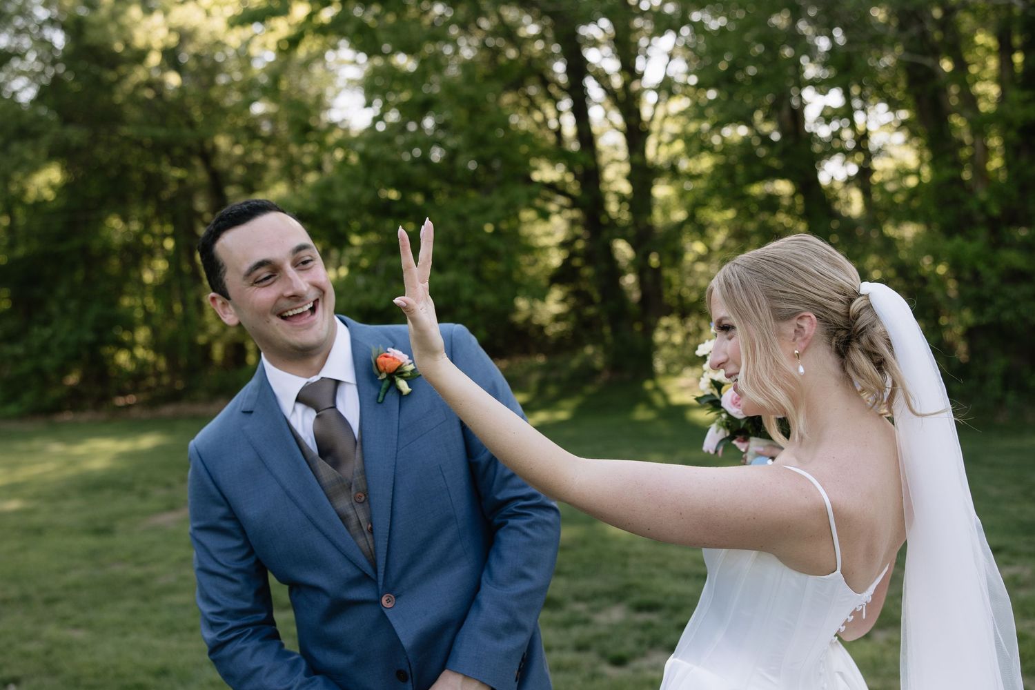 Bride playfully making bunny ears behind groom in blue suit during outdoor wedding photo session.