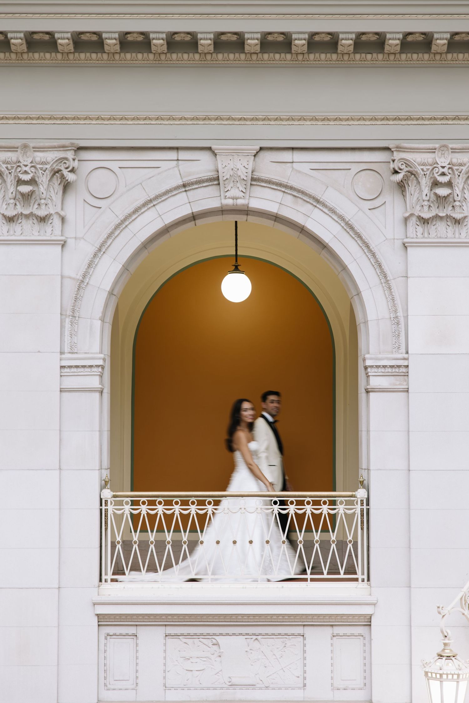 Bride and groom stand on ornate balcony framed by elegant white arch in classic wedding venue.