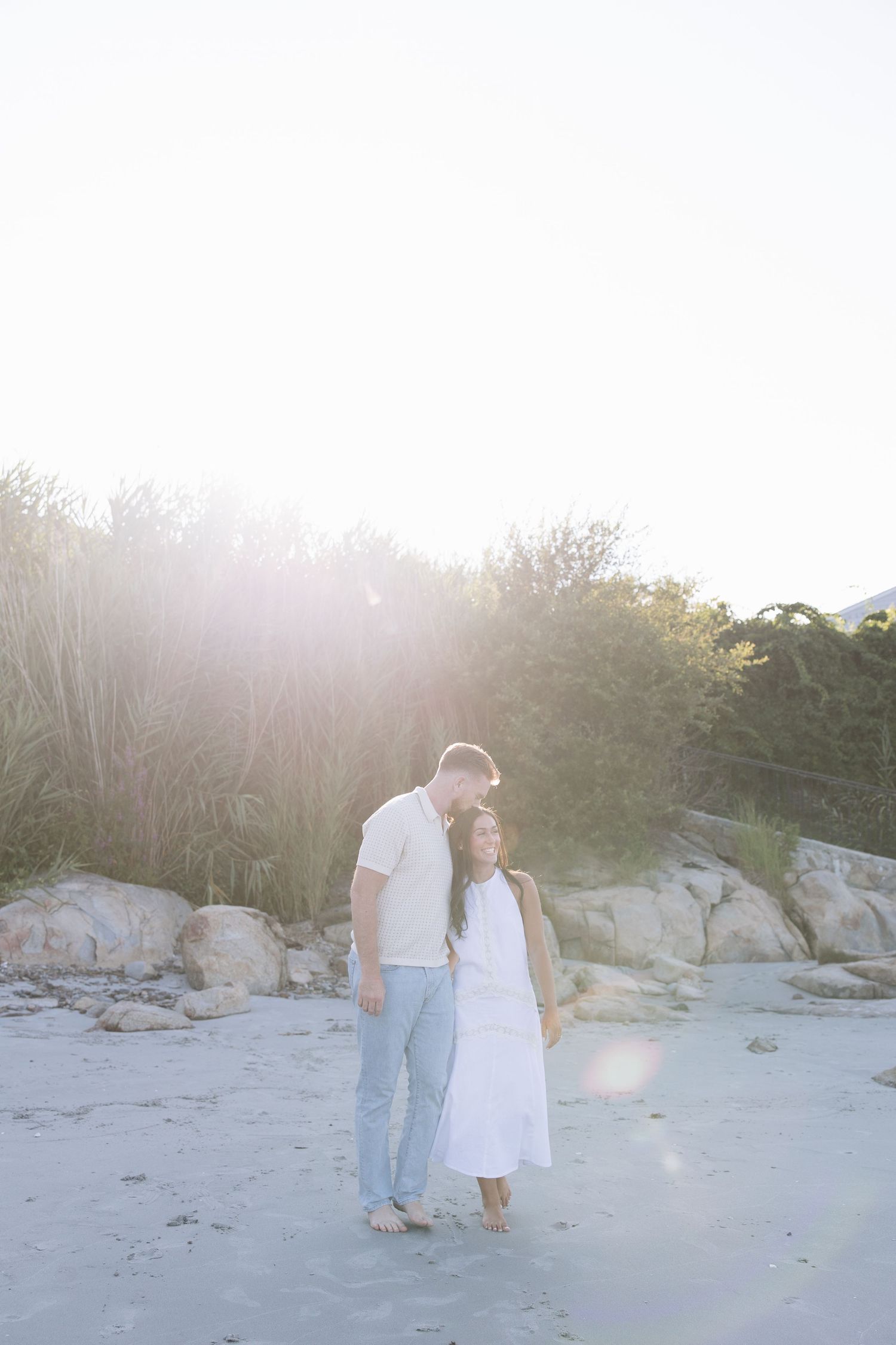 Couple embracing on a sunlit beach with golden lens flare, rocks, and lush greenery in the background.