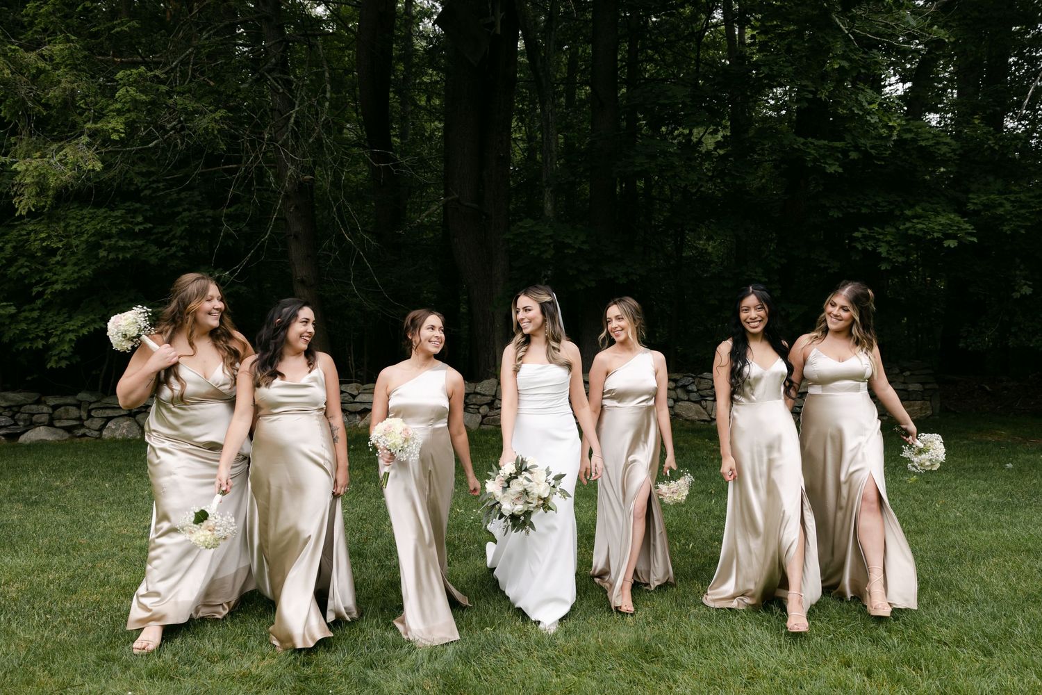 Bridesmaids in light colored dresses sit together on grass during outdoor wedding photoshoot.