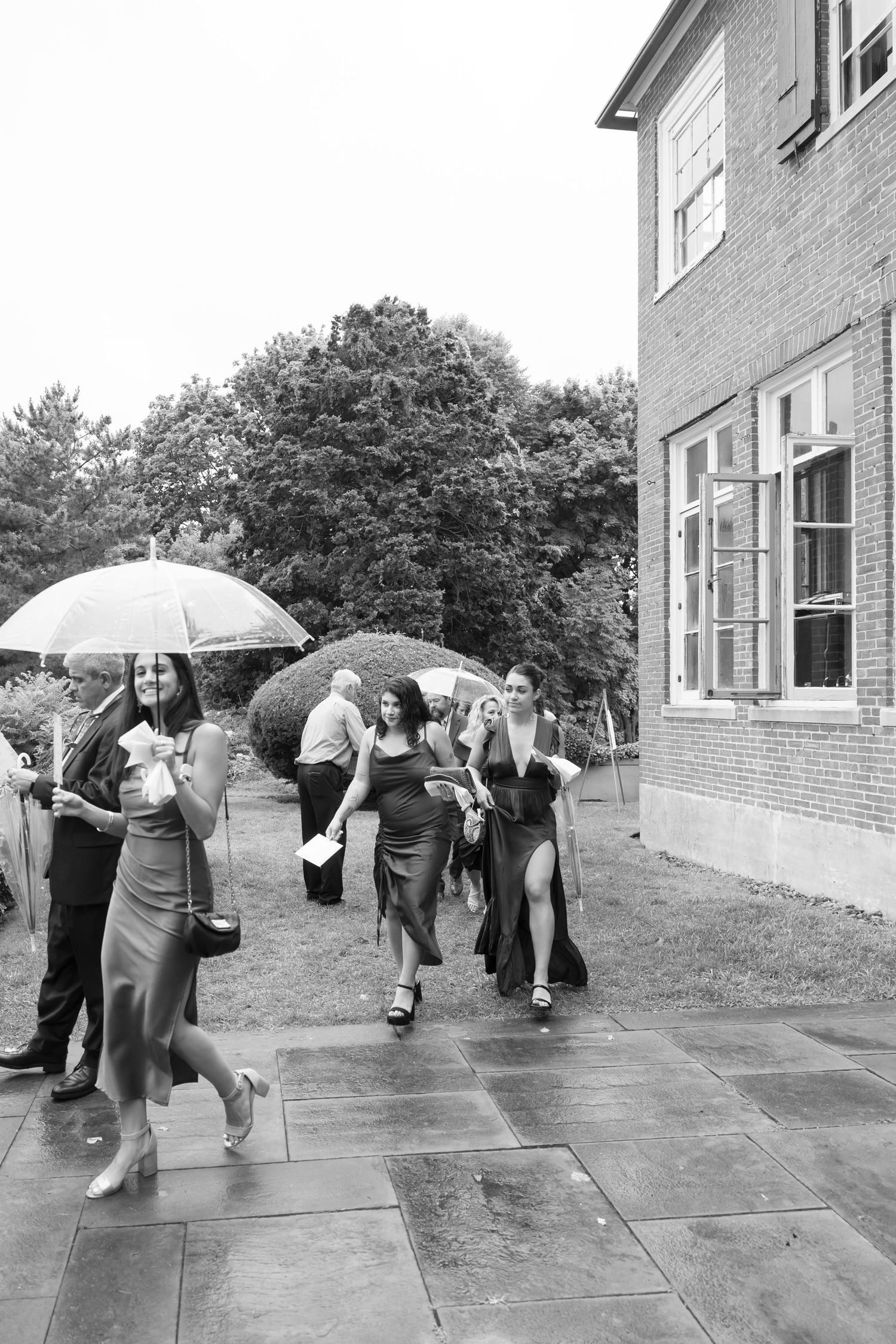 Wedding party members walk with umbrellas along a wet sidewalk outside a brick building.