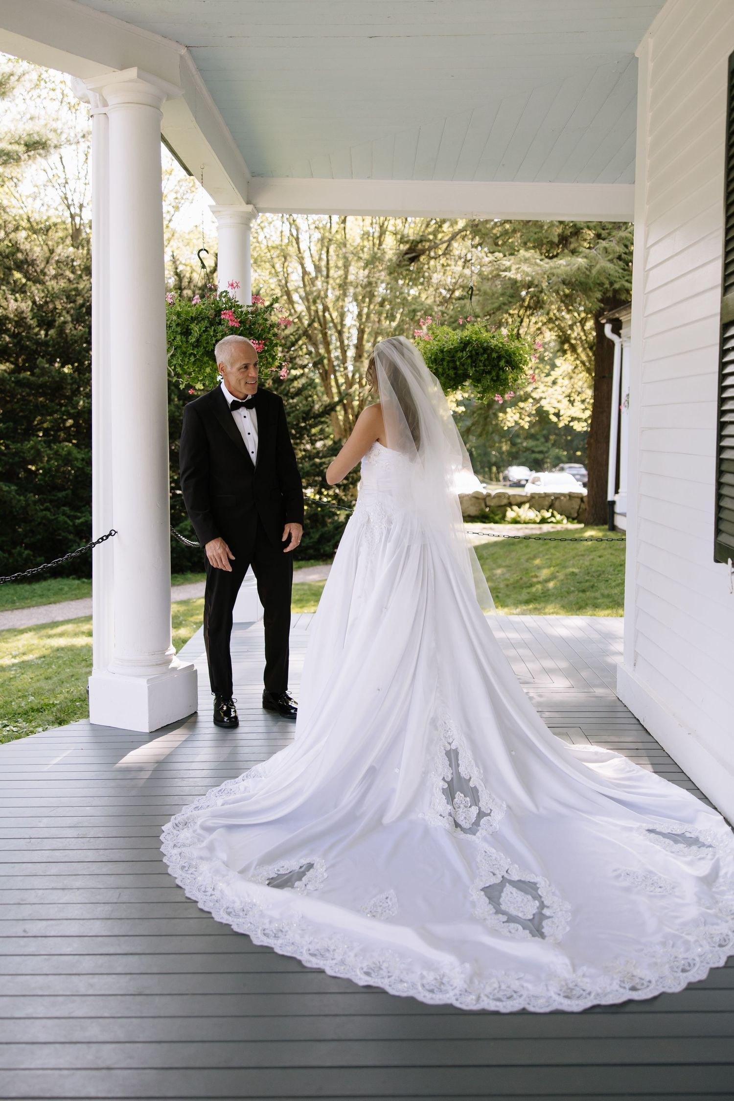 Bride in elaborate lace wedding gown with long train walks along white columned porch.