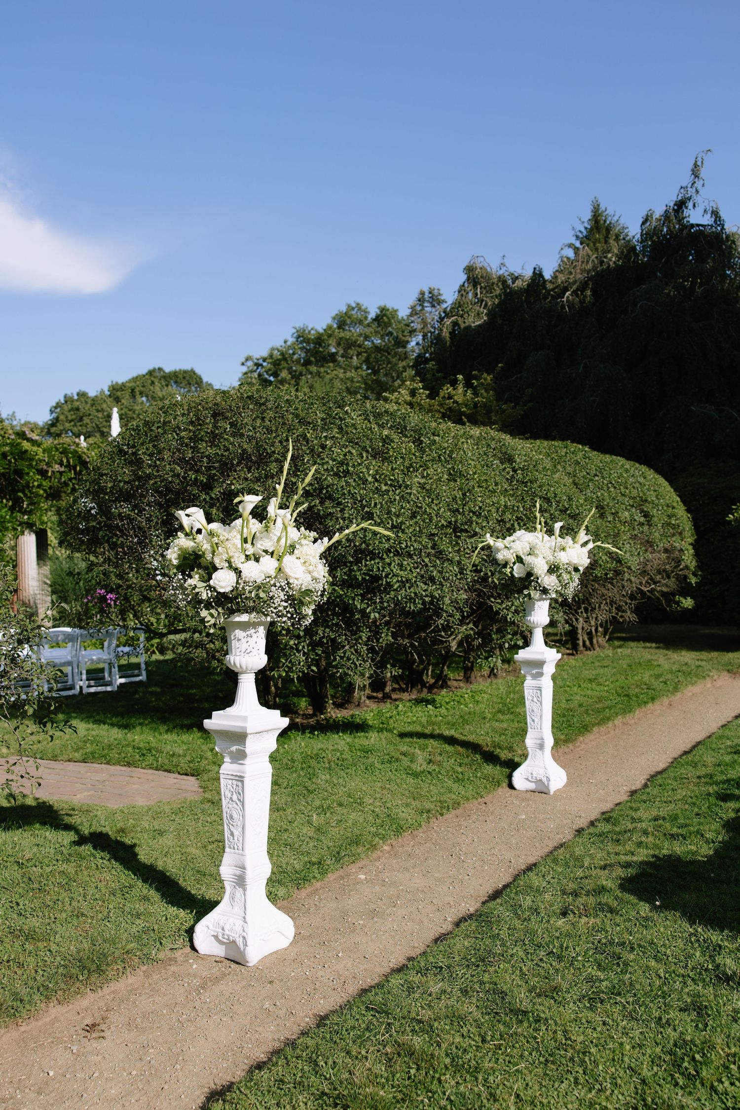 White floral arrangements on pedestals line a garden path for a wedding ceremony.