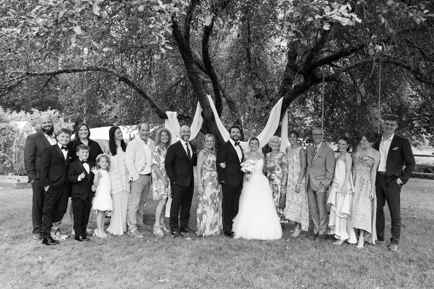 Black and white wedding photo of a large group posing under a tree outdoors on a sunny day.