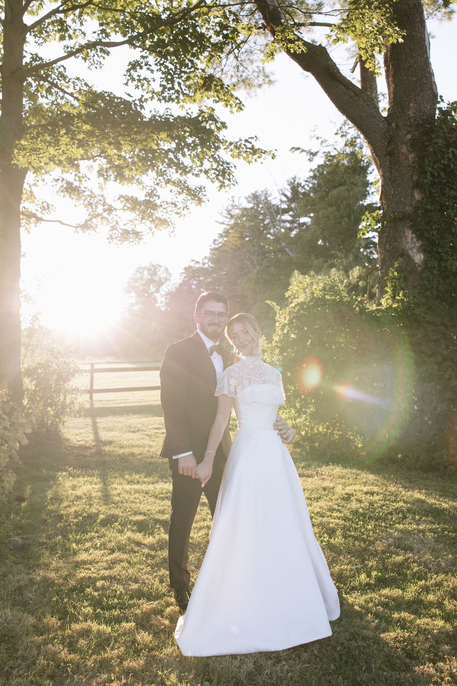 Bride and groom portrait in golden sunset light beneath large trees in a lush green outdoor setting.