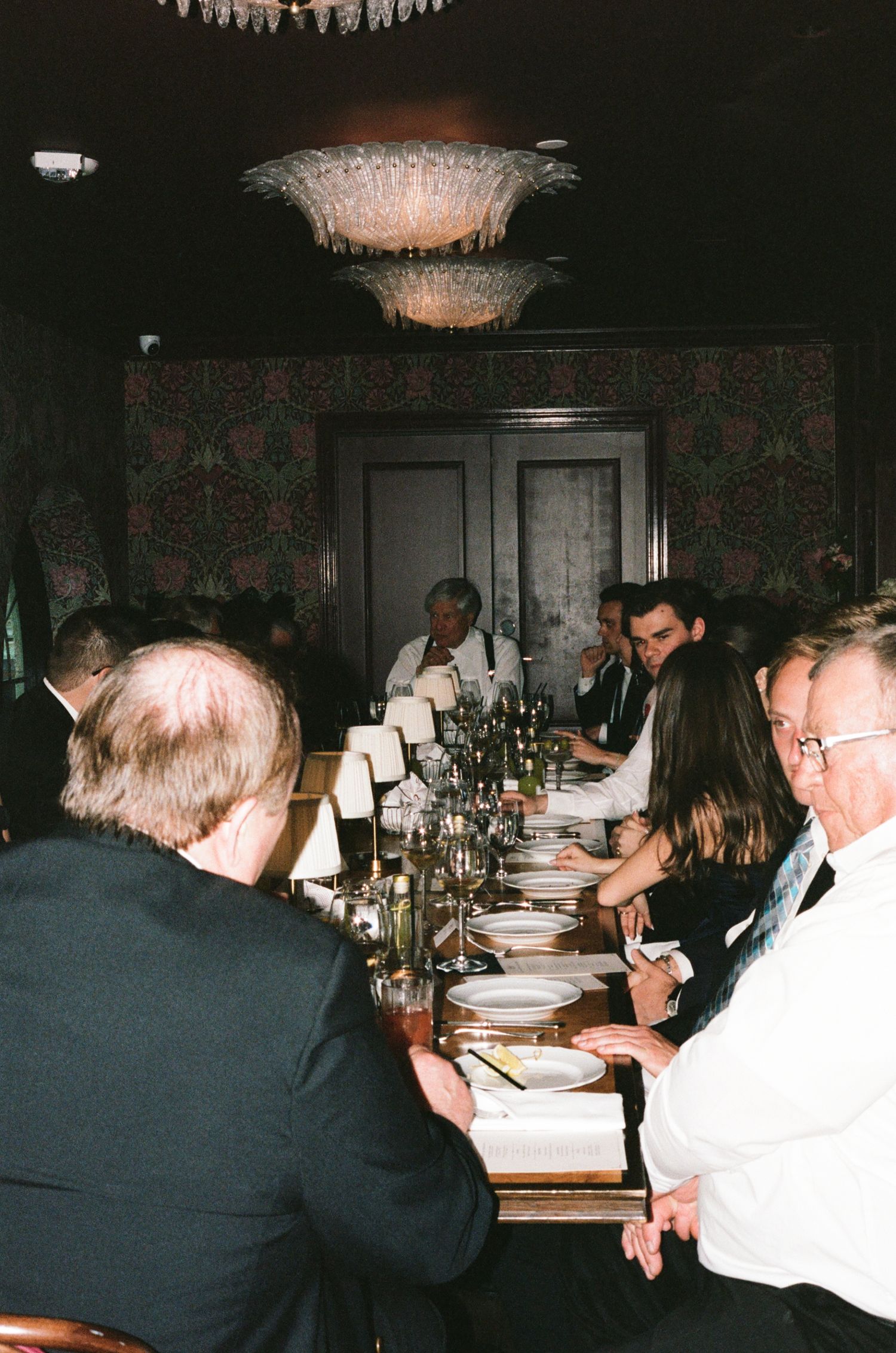 Guests seated at a long formal dining table in an elegant restaurant with chandelier lighting and floral wallpaper.