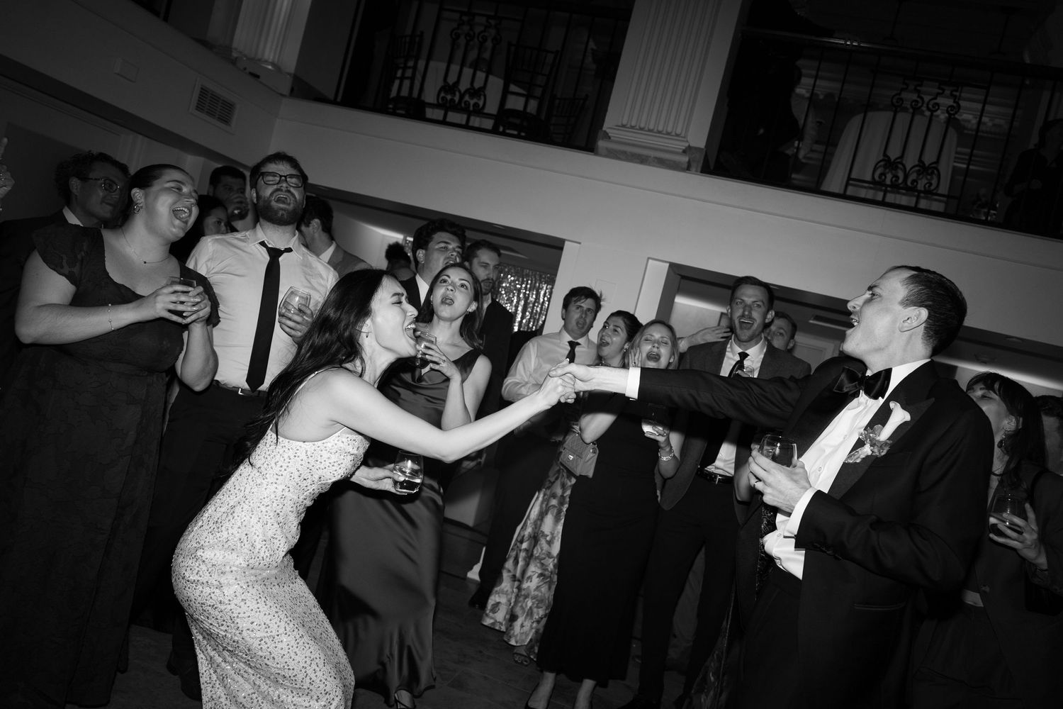 Black and white photo of bride and groom dancing joyfully with wedding guests at elegant indoor reception.