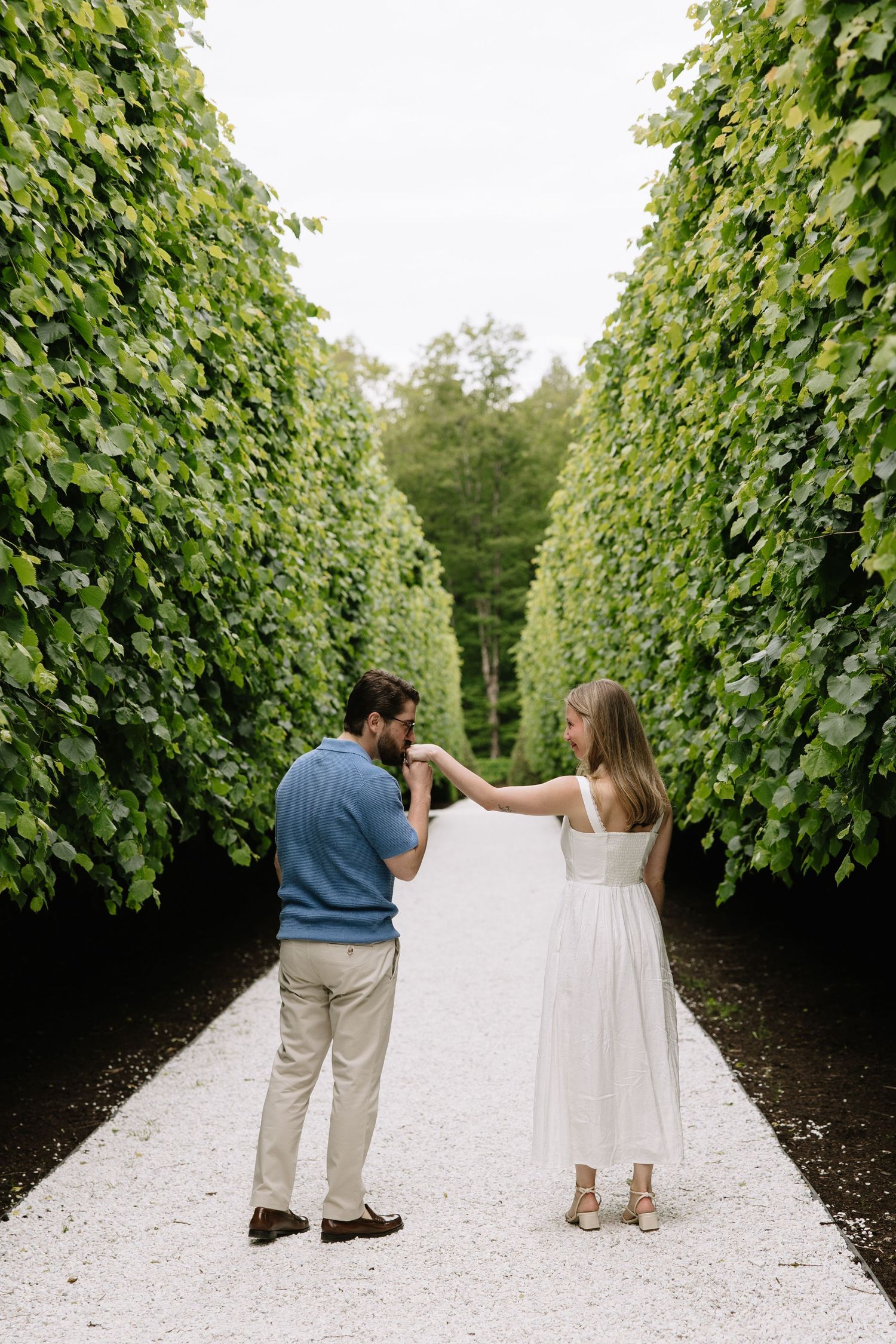 Couple holding hands on a white gravel path lined with tall green hedges in a romantic garden setting.