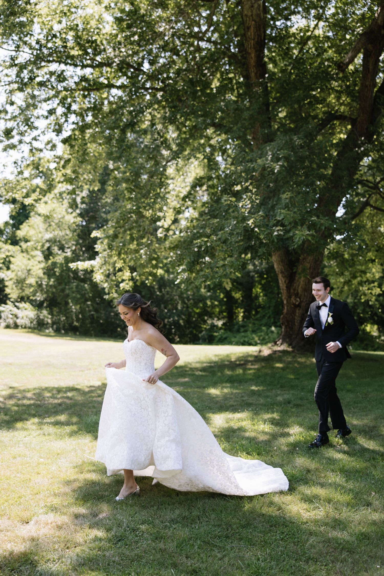 Bride in white gown and groom in black tuxedo walking on green grass beneath large trees on wedding day.