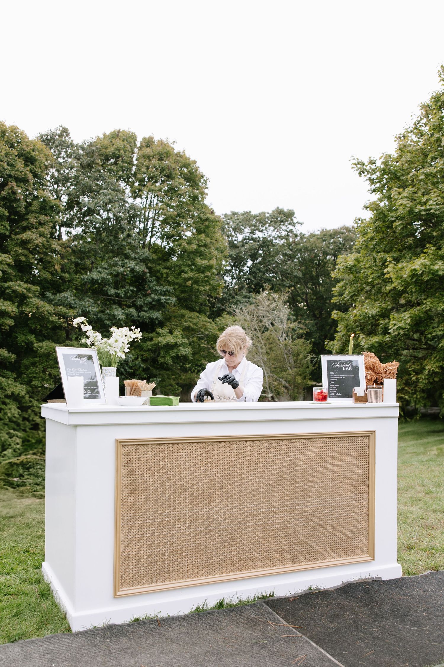 Elegant white outdoor wedding bar with burlap panels set up on a lawn.