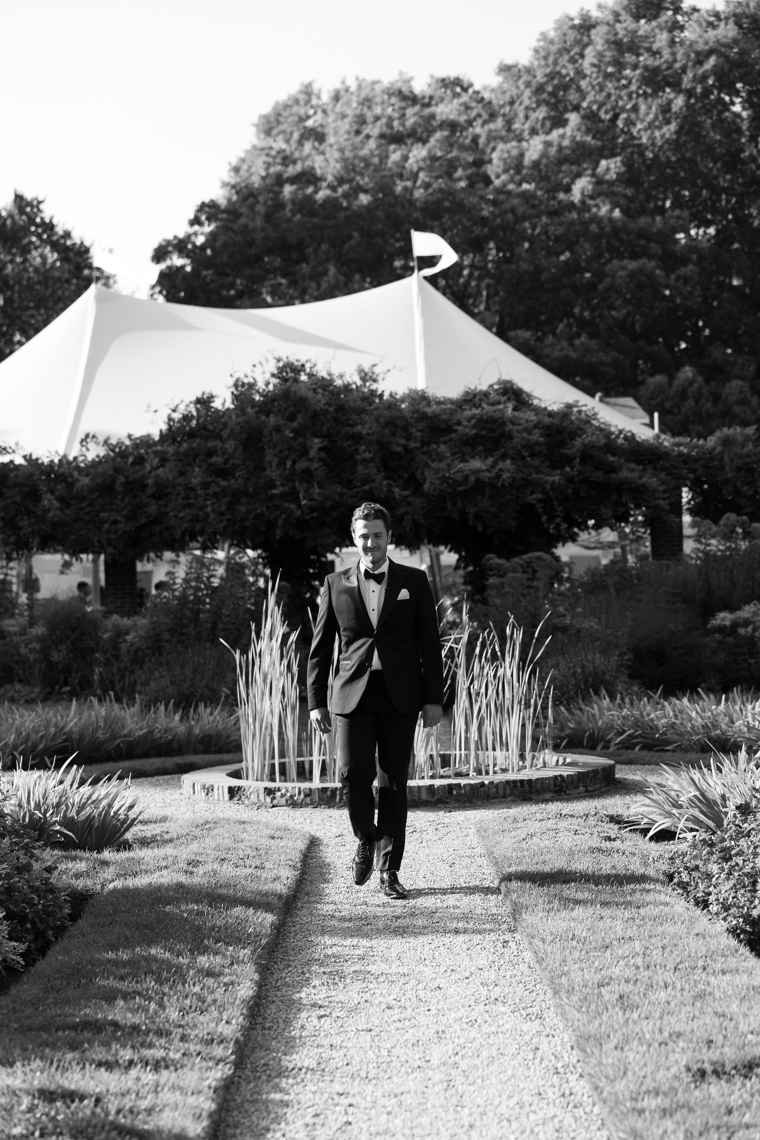 Black and white photo of a couple walking along a garden path with white tents in the distance.