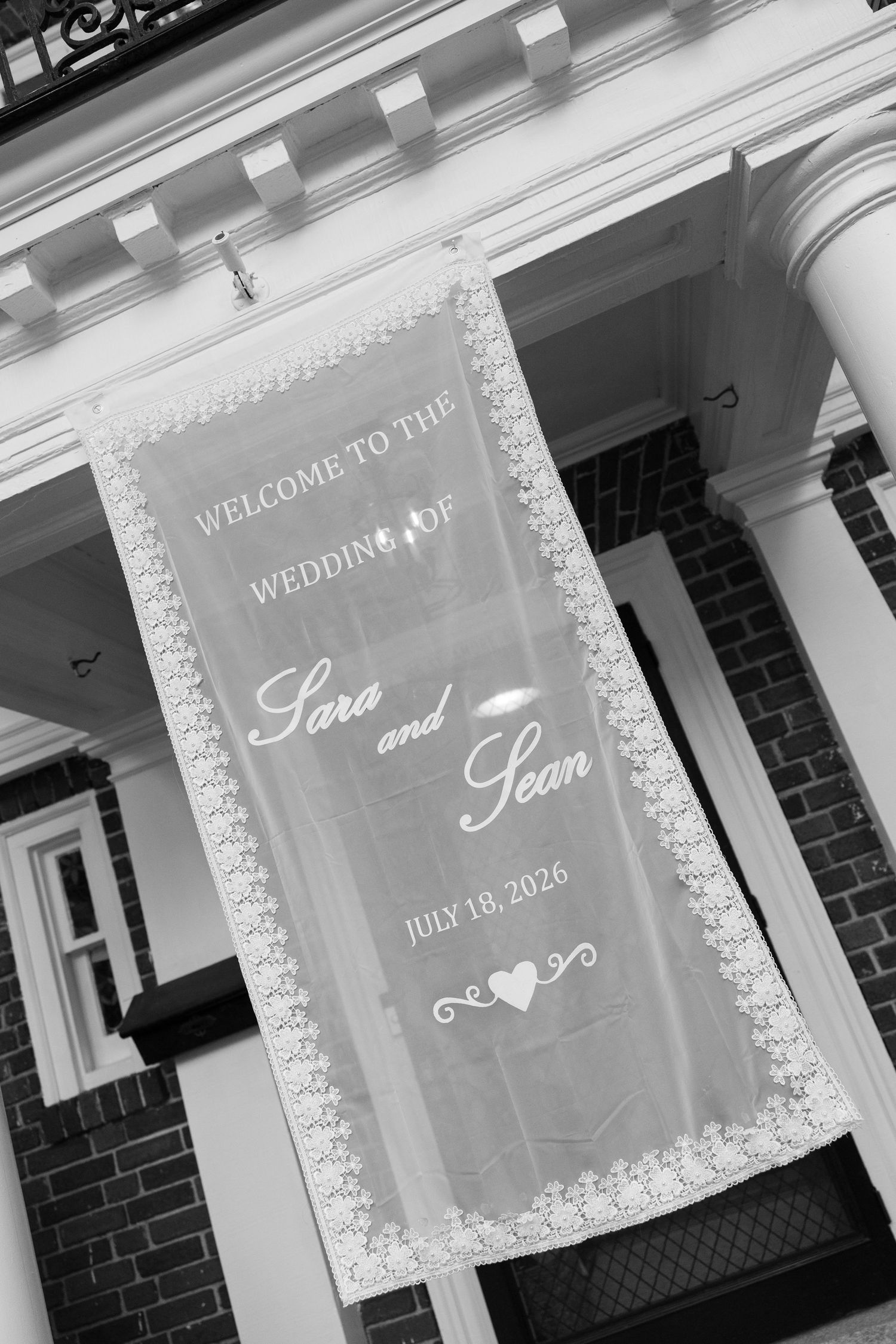 Black and white wedding welcome banner reading Sara and Sean July 18 2026 hanging on classic brick building columns.