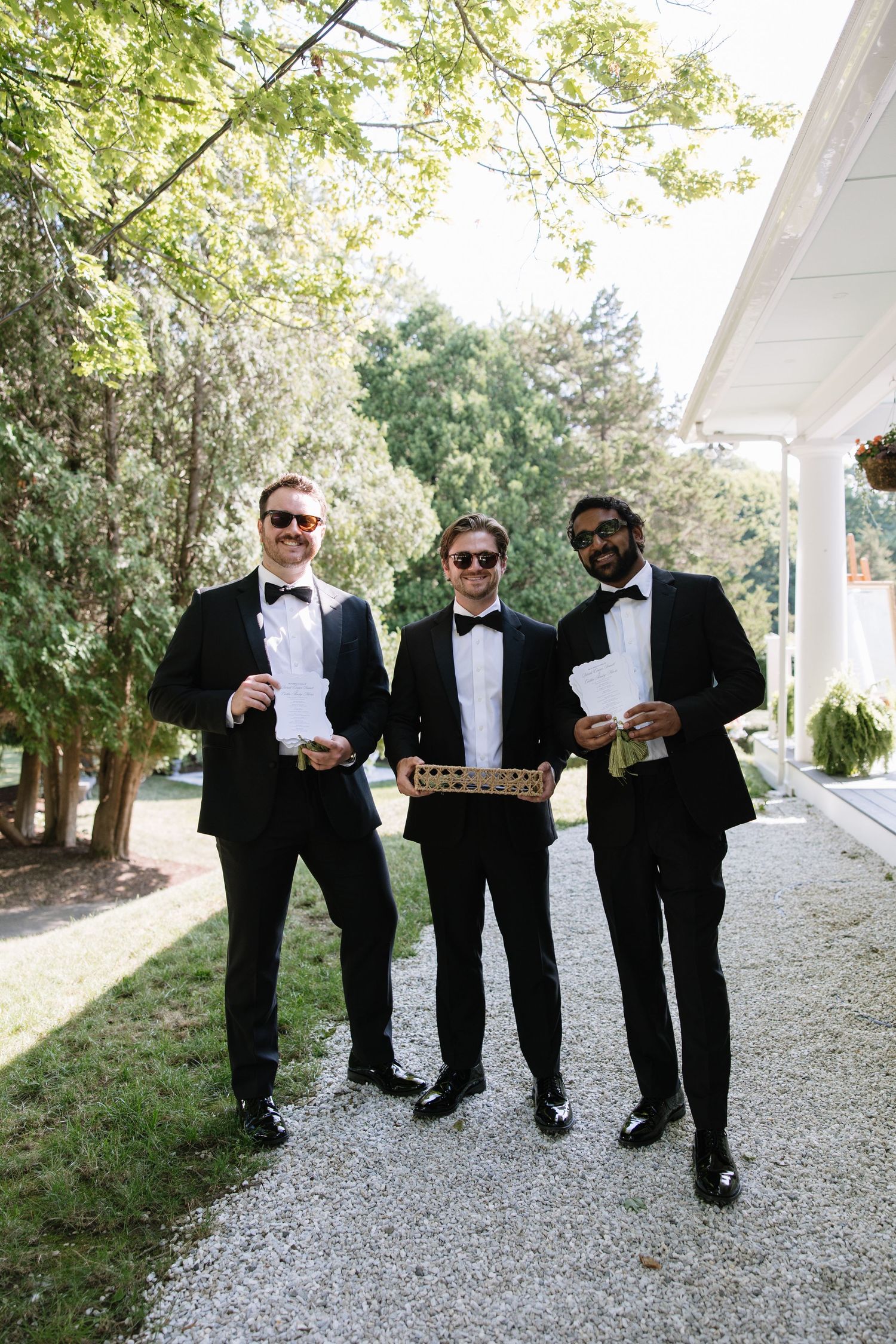 Three groomsmen in black tuxedos and sunglasses holding wedding programs outdoors on a gravel path.