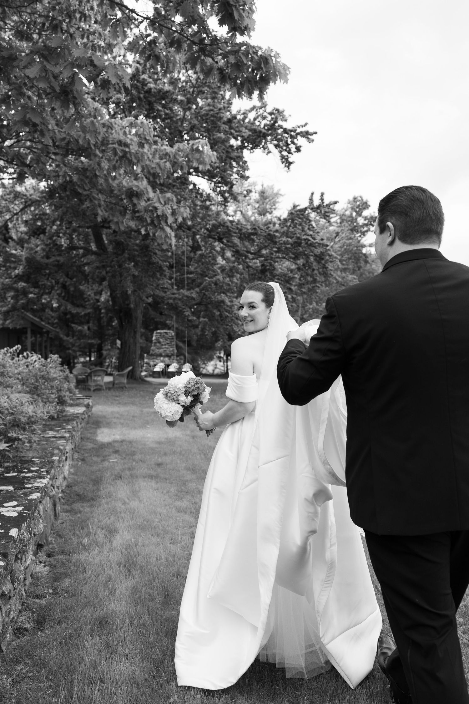 Black and white photo of a smiling bride in white gown holding bouquet, with groom adjusting her veil outdoors.
