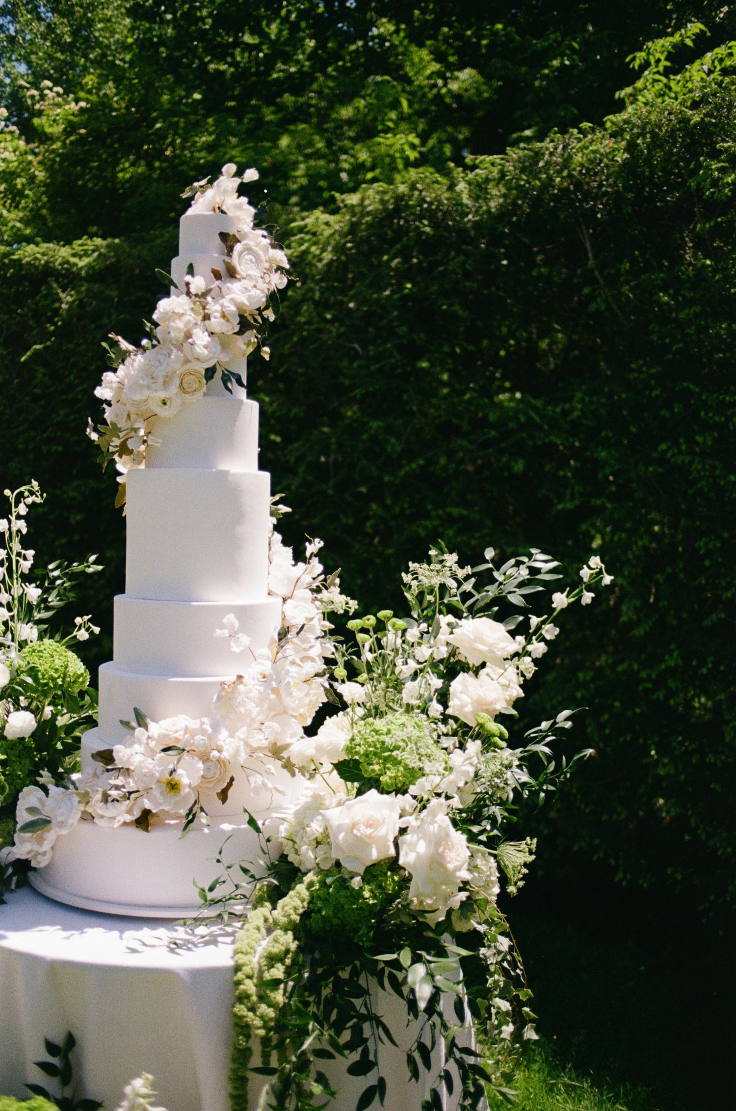 Elegant white wedding cake adorned with cascading white orchids and greenery against a garden backdrop.