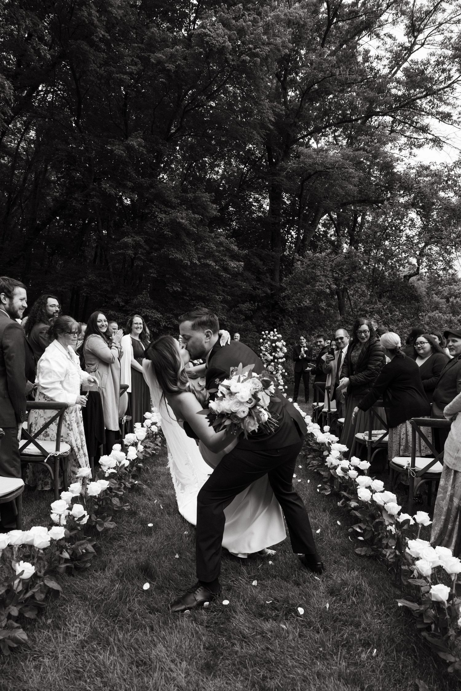Black and white wedding photo showing guests celebrating on a grassy hillside with floral decor.
