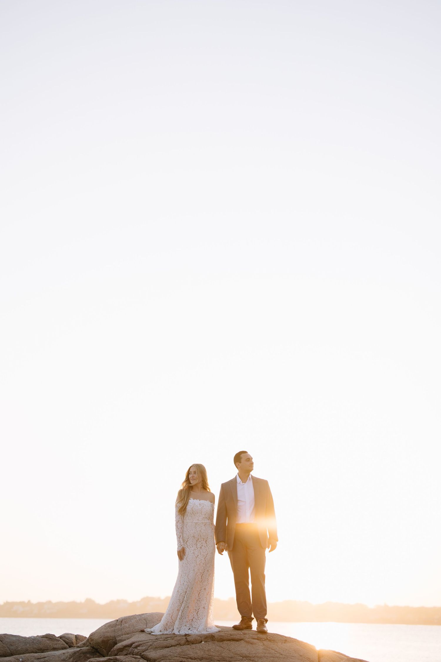 Two people standing together on rocks against a bright sunset sky.