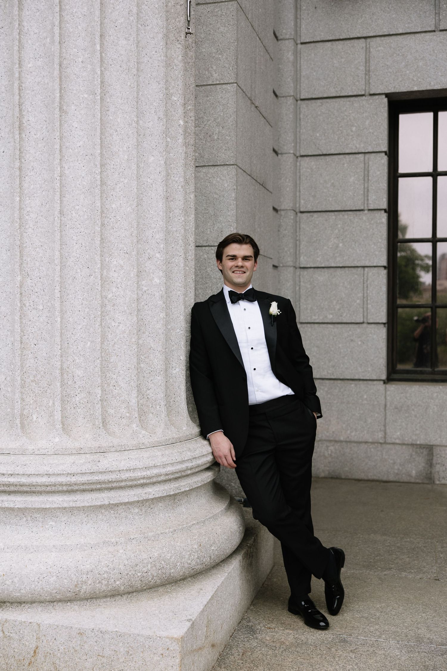 Groom in black tuxedo with bow tie leans against stone column at elegant venue.