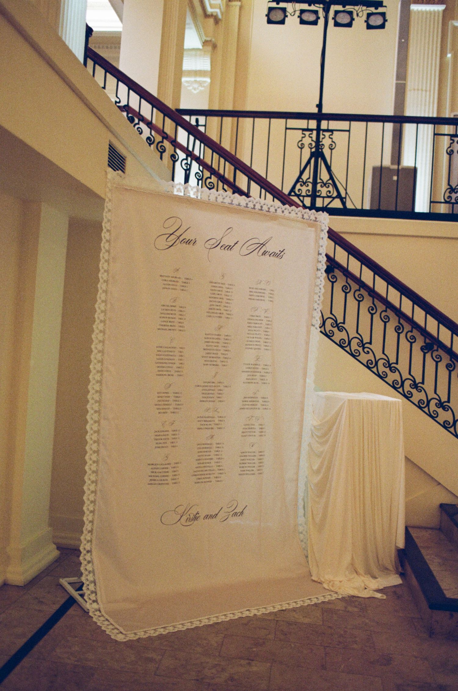 Elegant wedding seating chart displayed on a lace fabric banner near a grand staircase in a formal venue.