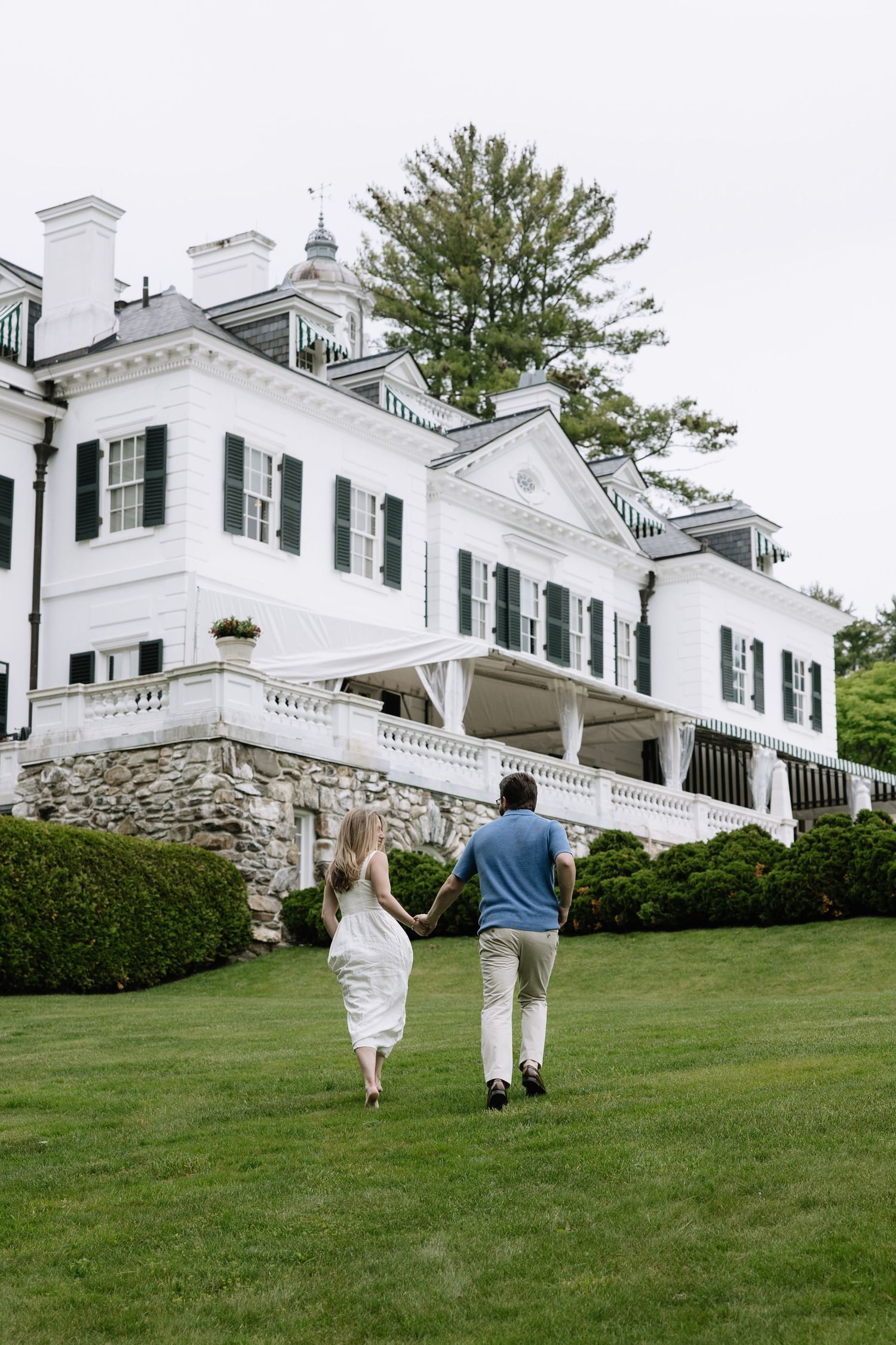 Couple running hand in hand across green lawn toward a classic white colonial mansion with dark shutters.