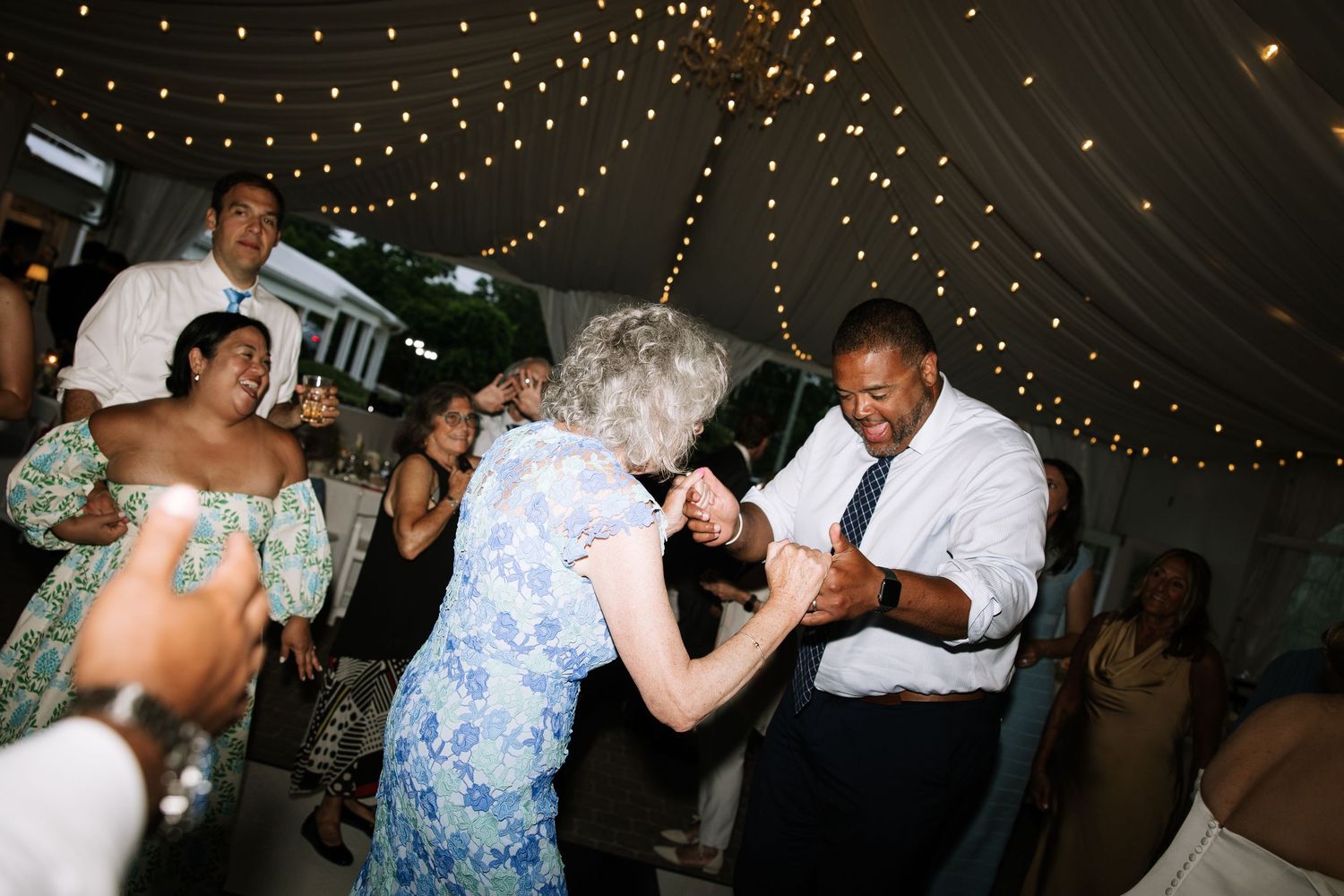 Elderly woman and man dancing joyfully at a wedding reception under twinkling string lights.