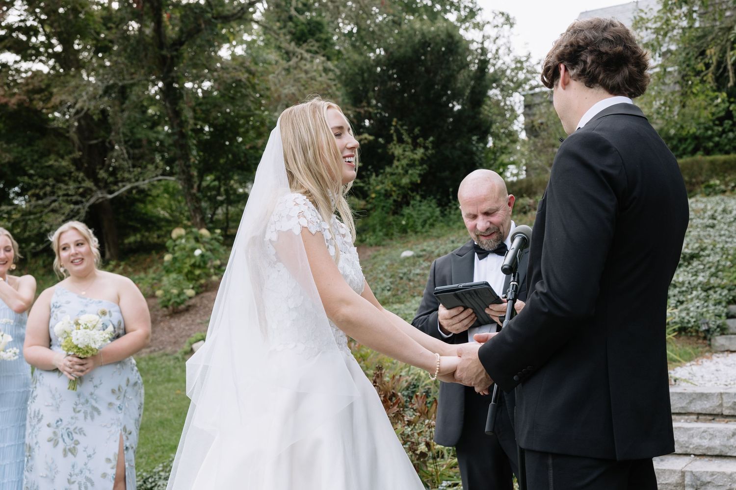 Bride in white lace gown laughing with groom in black tuxedo during outdoor wedding ceremony with officiant.