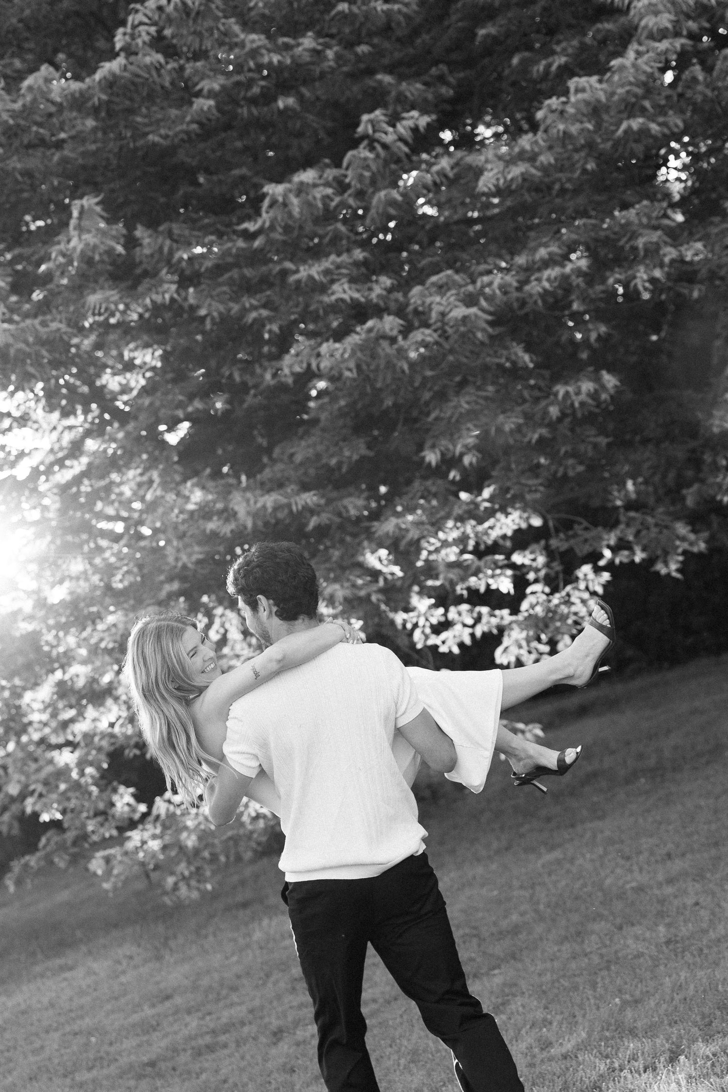 Black and white photo of a man lifting and spinning a woman outdoors in a sunlit park setting.