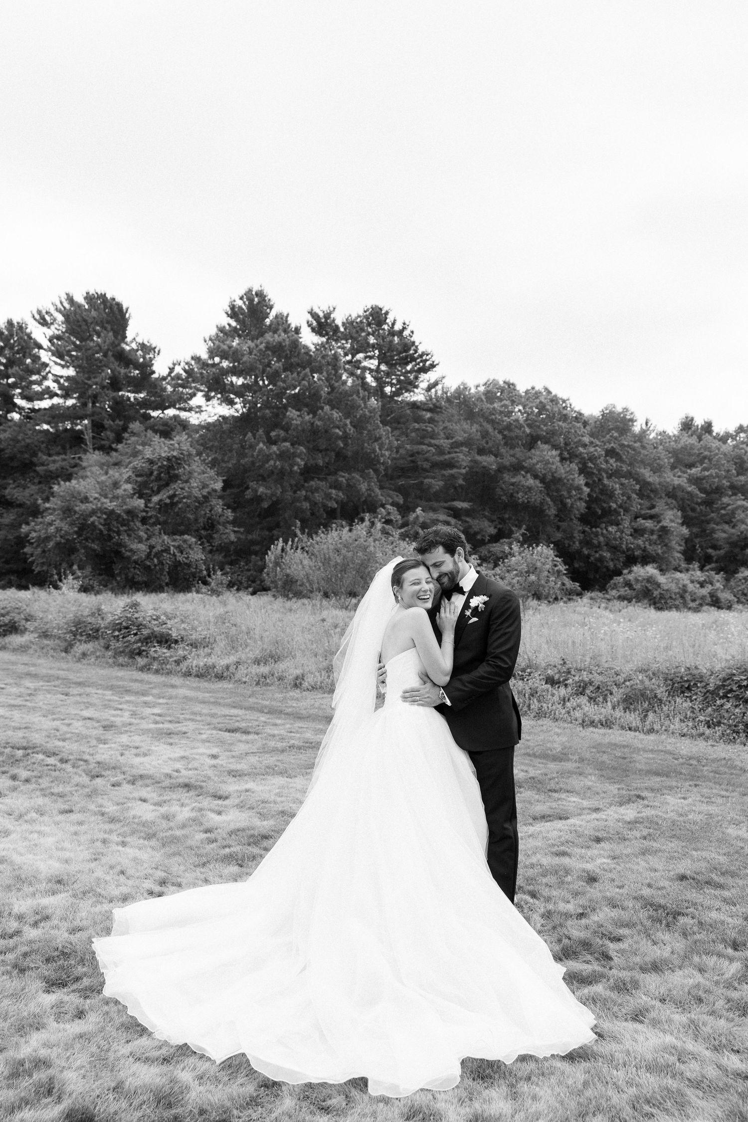 Black and white photo of bride and groom embracing in a field with trees in background on their wedding day.