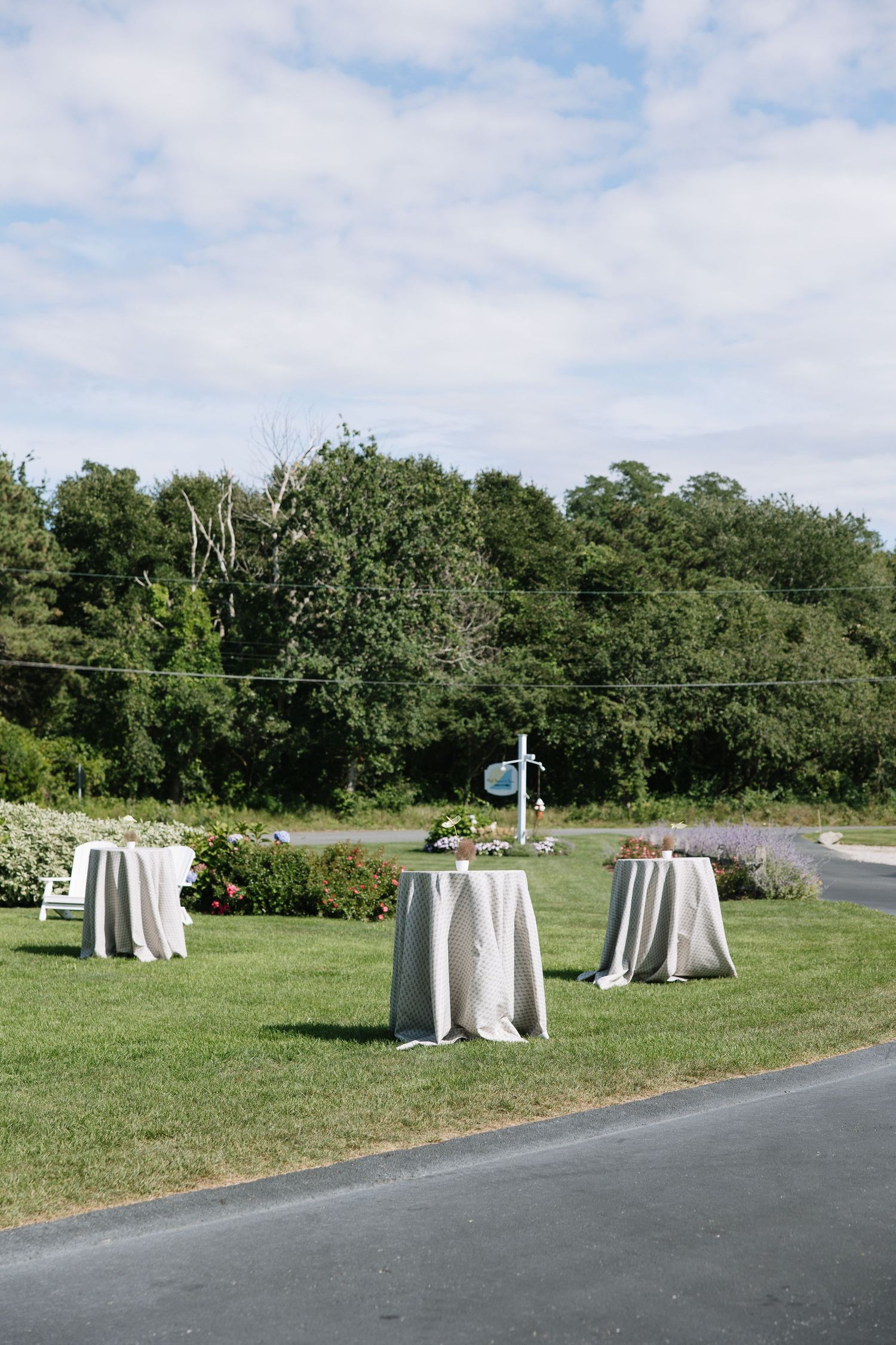 Elegant outdoor cocktail hour setup with linen-draped high-top tables on green lawn under blue sky.