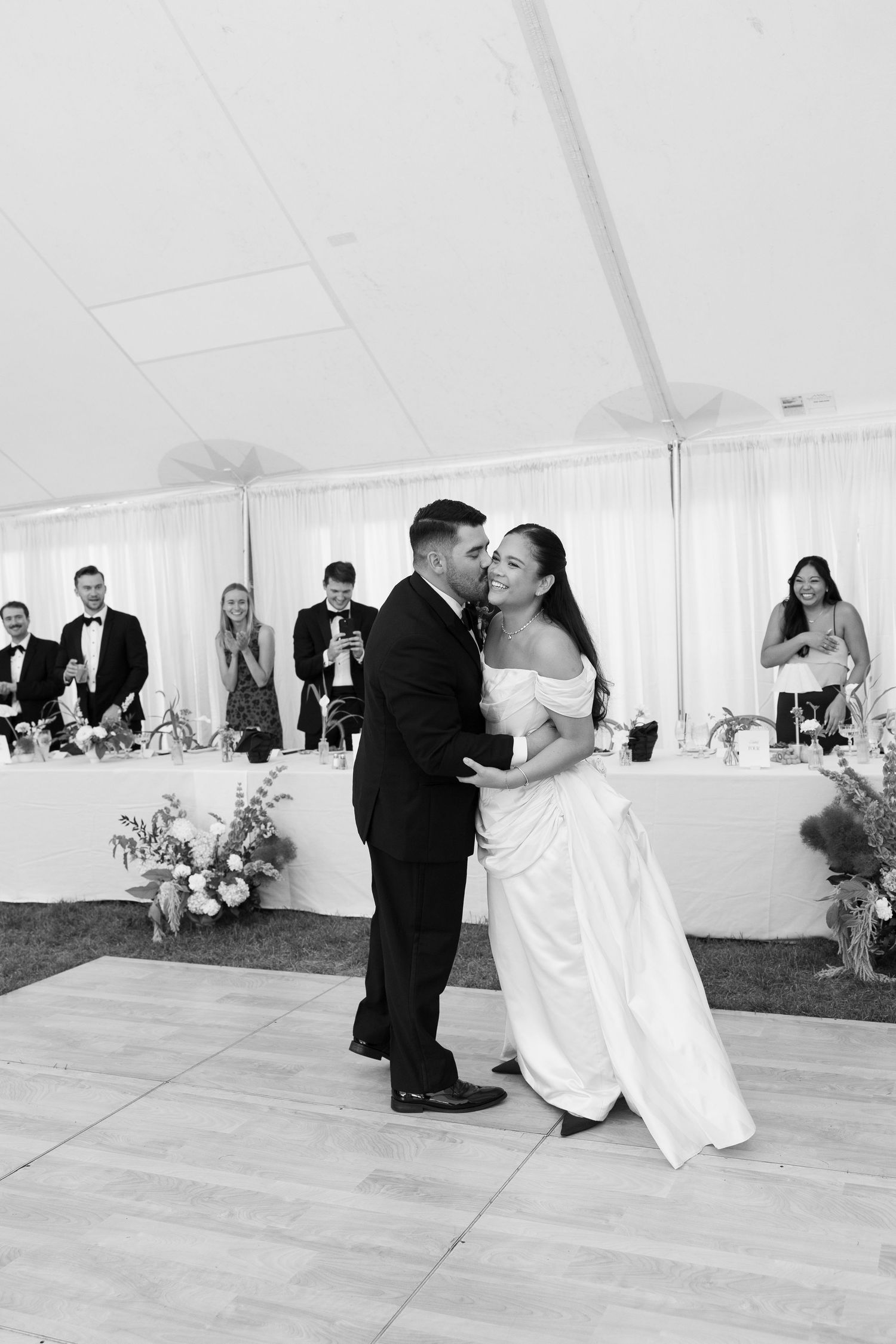 Bride and groom share a romantic first dance kiss in a white tent reception in black and white.