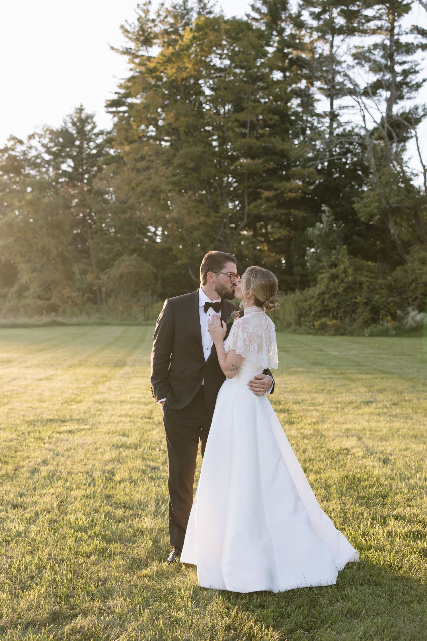 A bride and groom share a kiss on a sunlit lawn with tall trees in the background during their outdoor wedding.