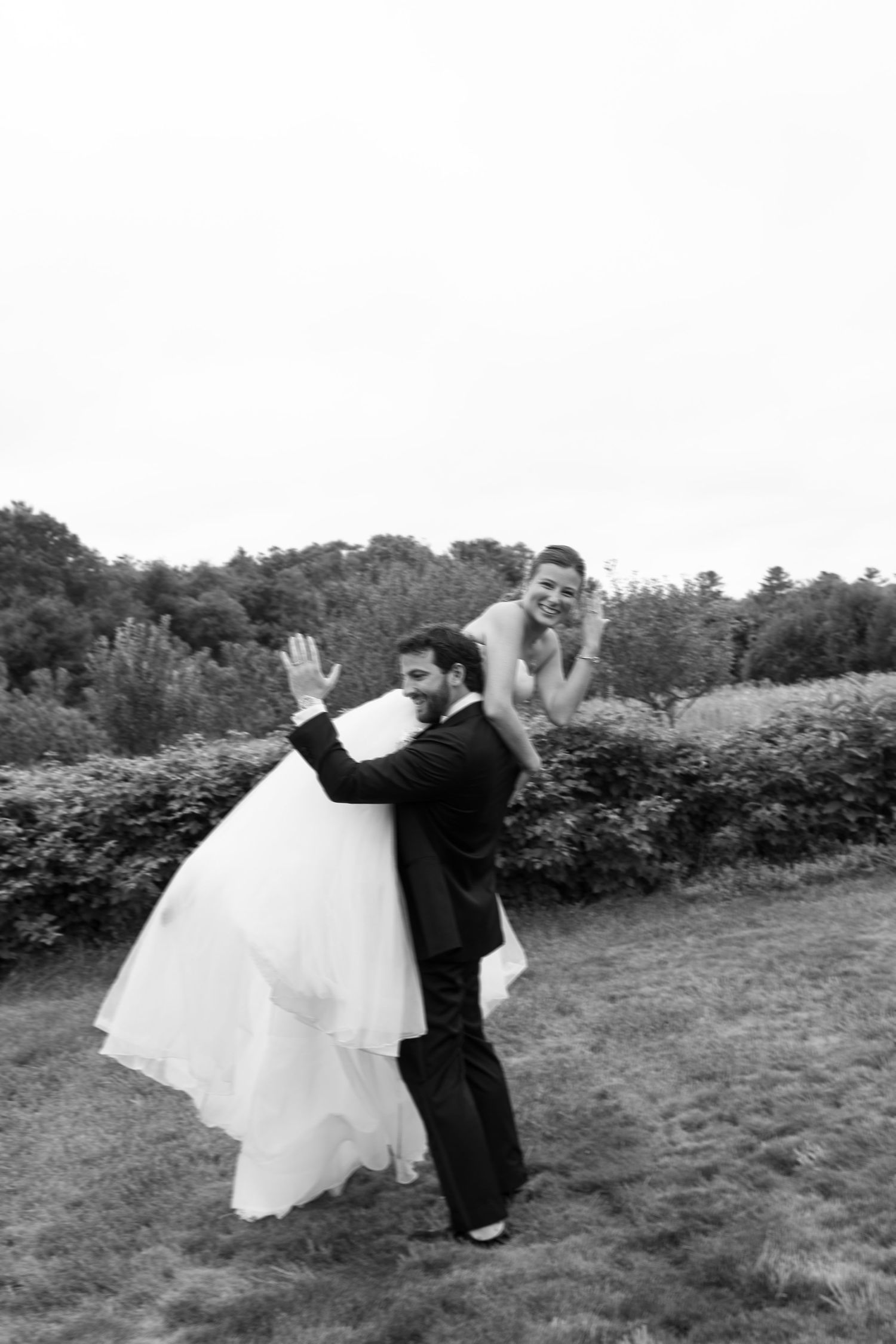 Groom playfully carrying bride in wedding dress outdoors in black and white photo.