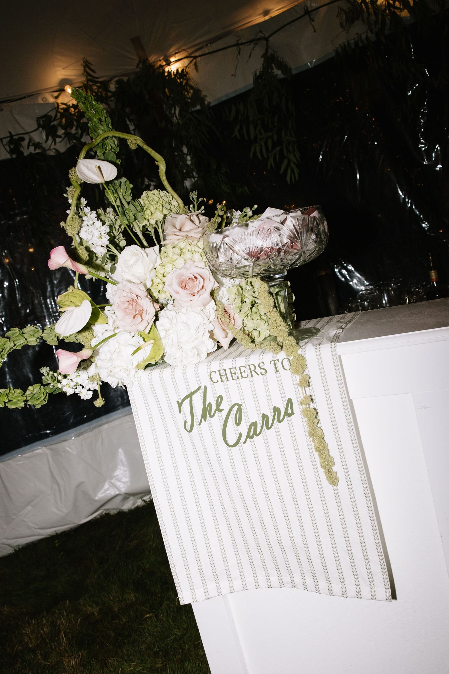 Wedding bar sign reading Cheers to The Carrs adorned with pink roses and white floral arrangement at reception.