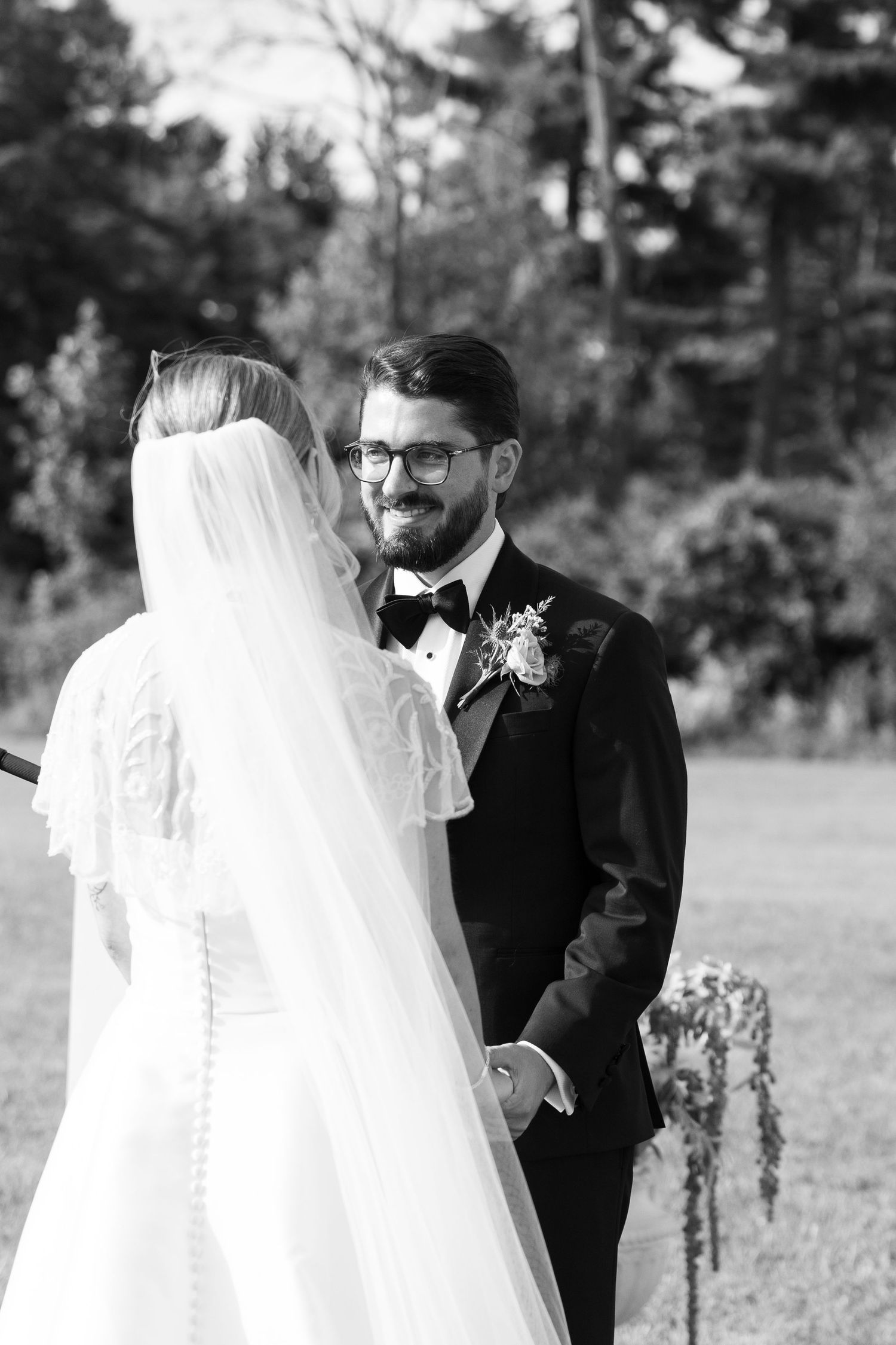 Black and white photo of a groom in a tuxedo smiling at his bride in a wedding veil during an outdoor ceremony.