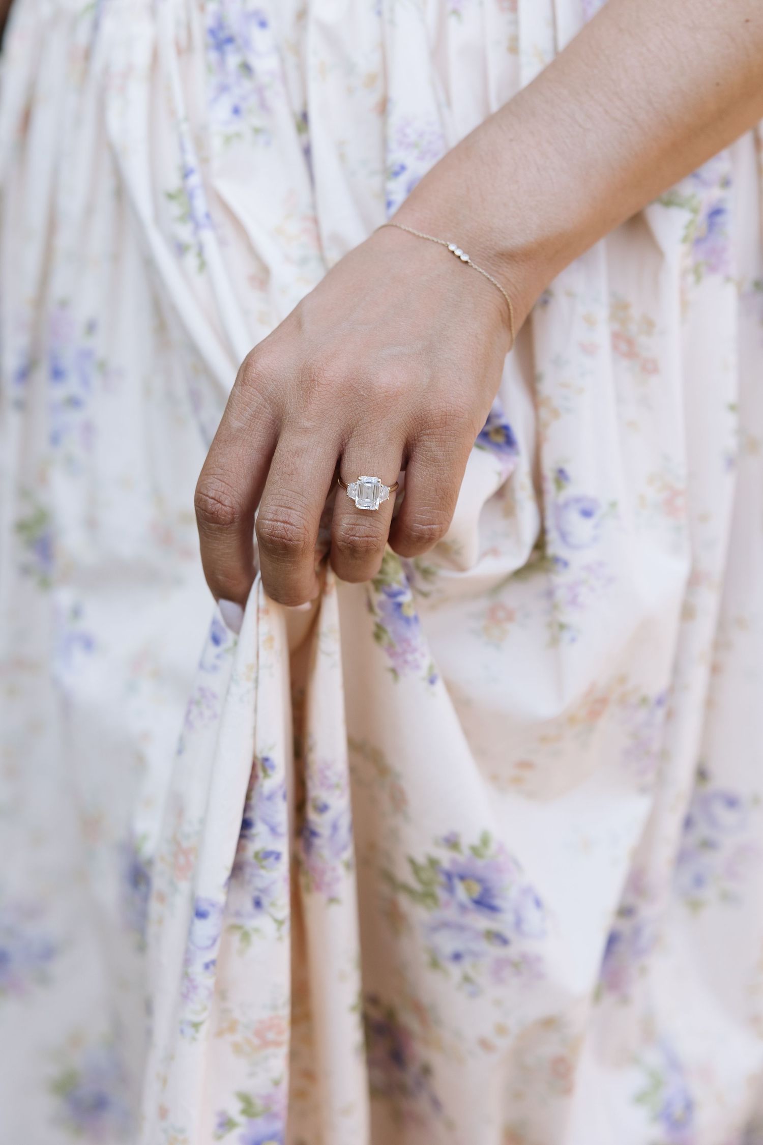 Close-up of a hand wearing an emerald-cut diamond engagement ring, holding a floral dress.
