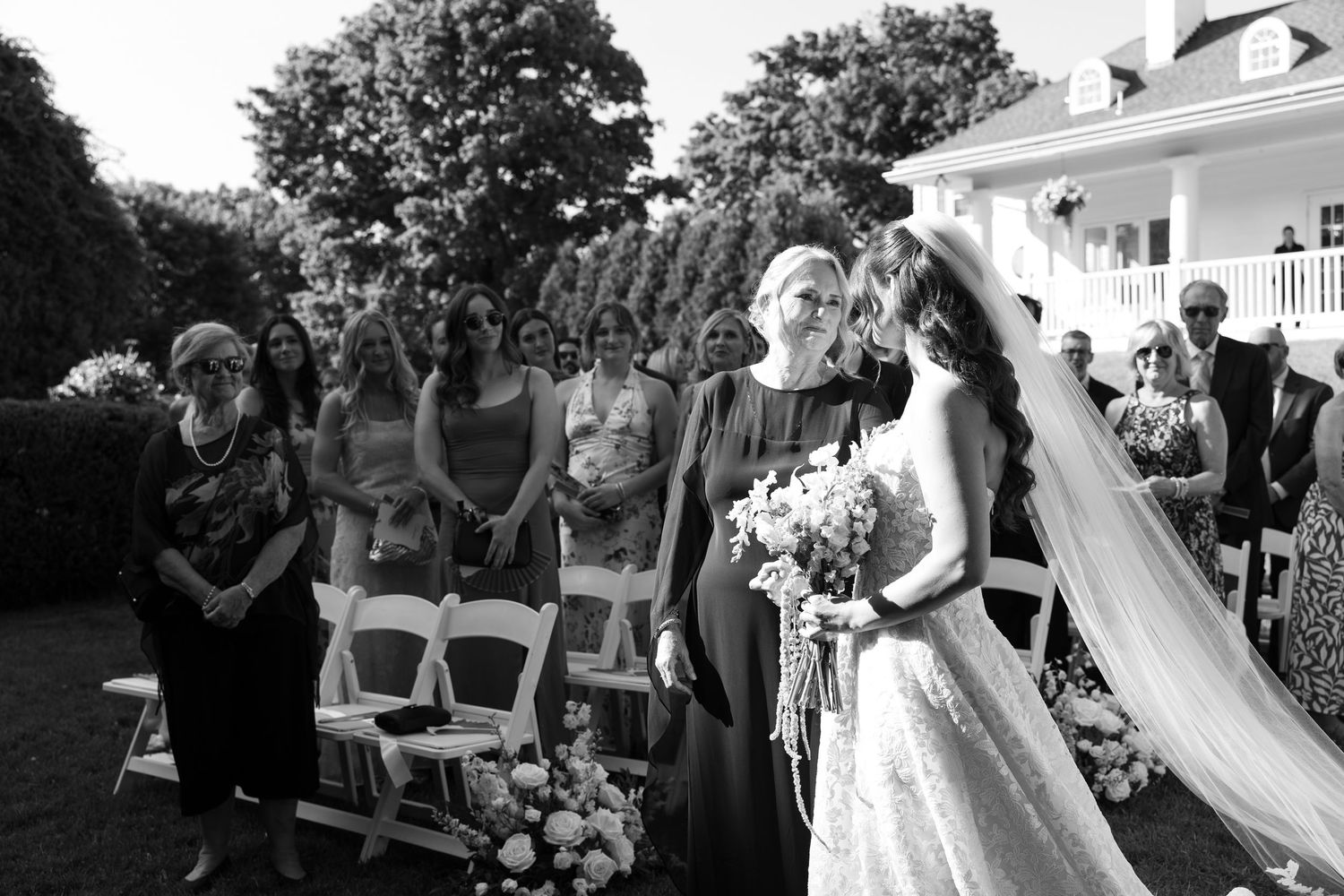 Black and white outdoor wedding ceremony with bride holding bouquet, guests seated on white chairs, and elegant venue backdrop.