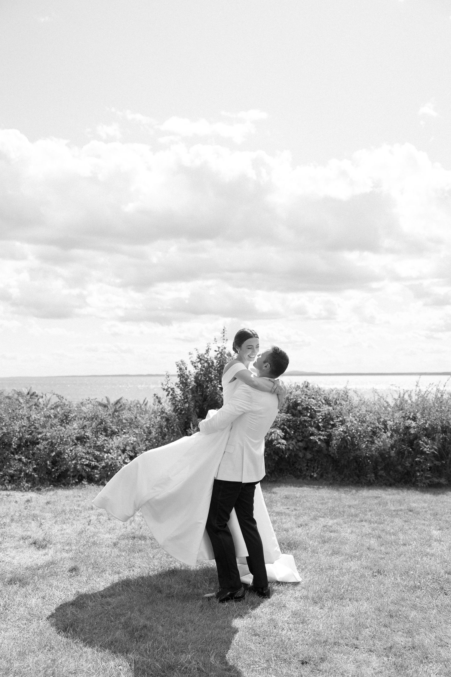 Black and white photo of groom lifting bride in wedding dress near waterfront with cloudy sky.