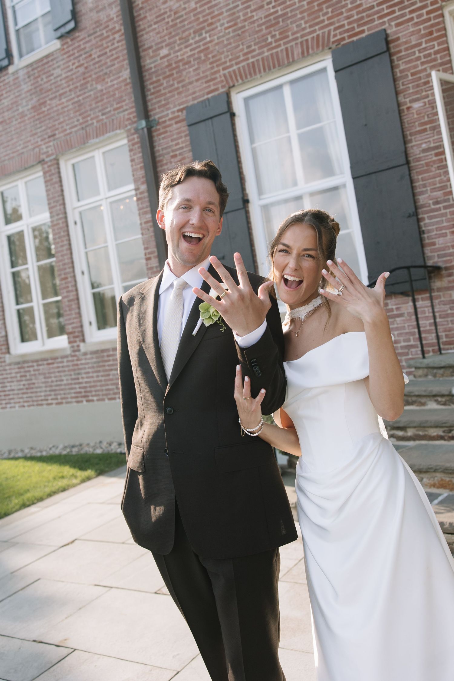 Joyful newlyweds showing off wedding rings outside brick building on their wedding day.