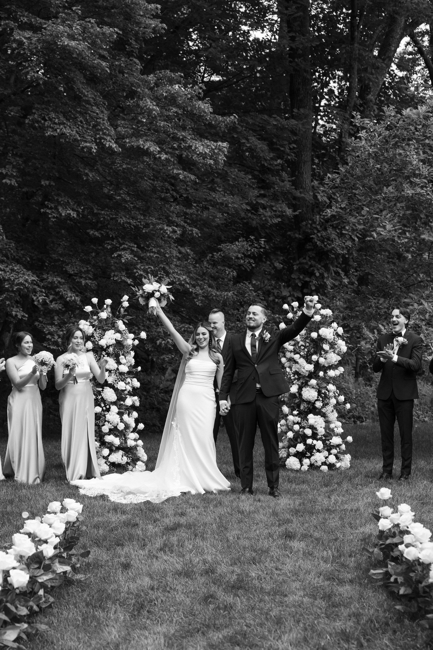 Wedding party poses for photos on grassy hill with white floral arrangements and trees in background.