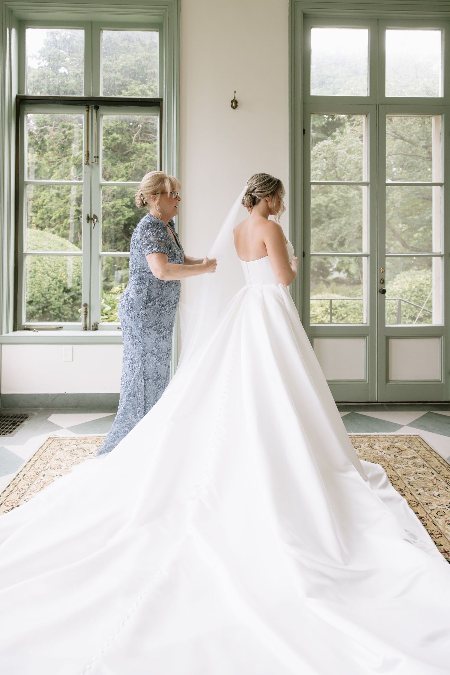 Wedding dress with flowing train being adjusted in a sunlit room with green windows.