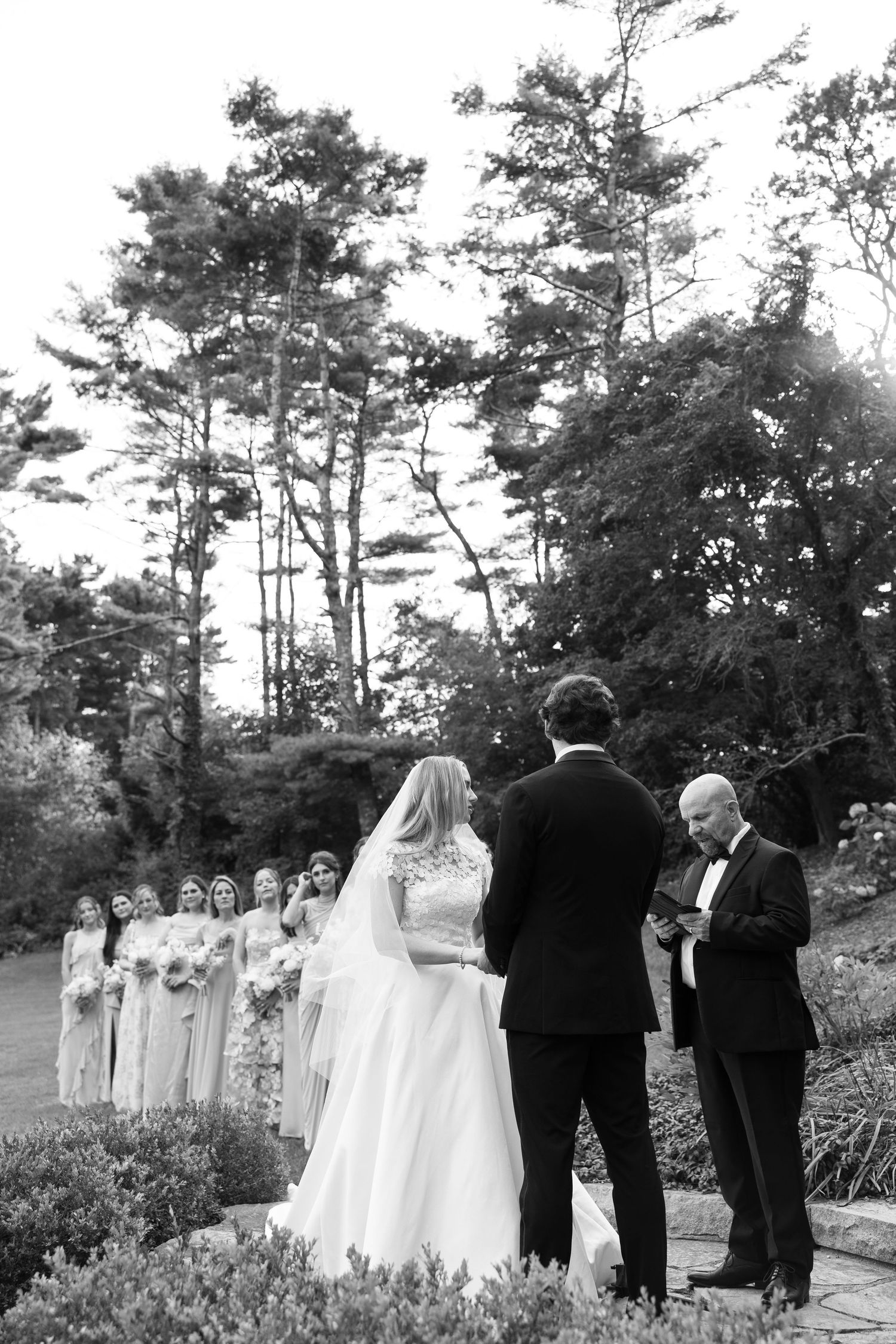 An outdoor wedding ceremony takes place among tall pine trees with bridesmaids in soft blue dresses looking on.