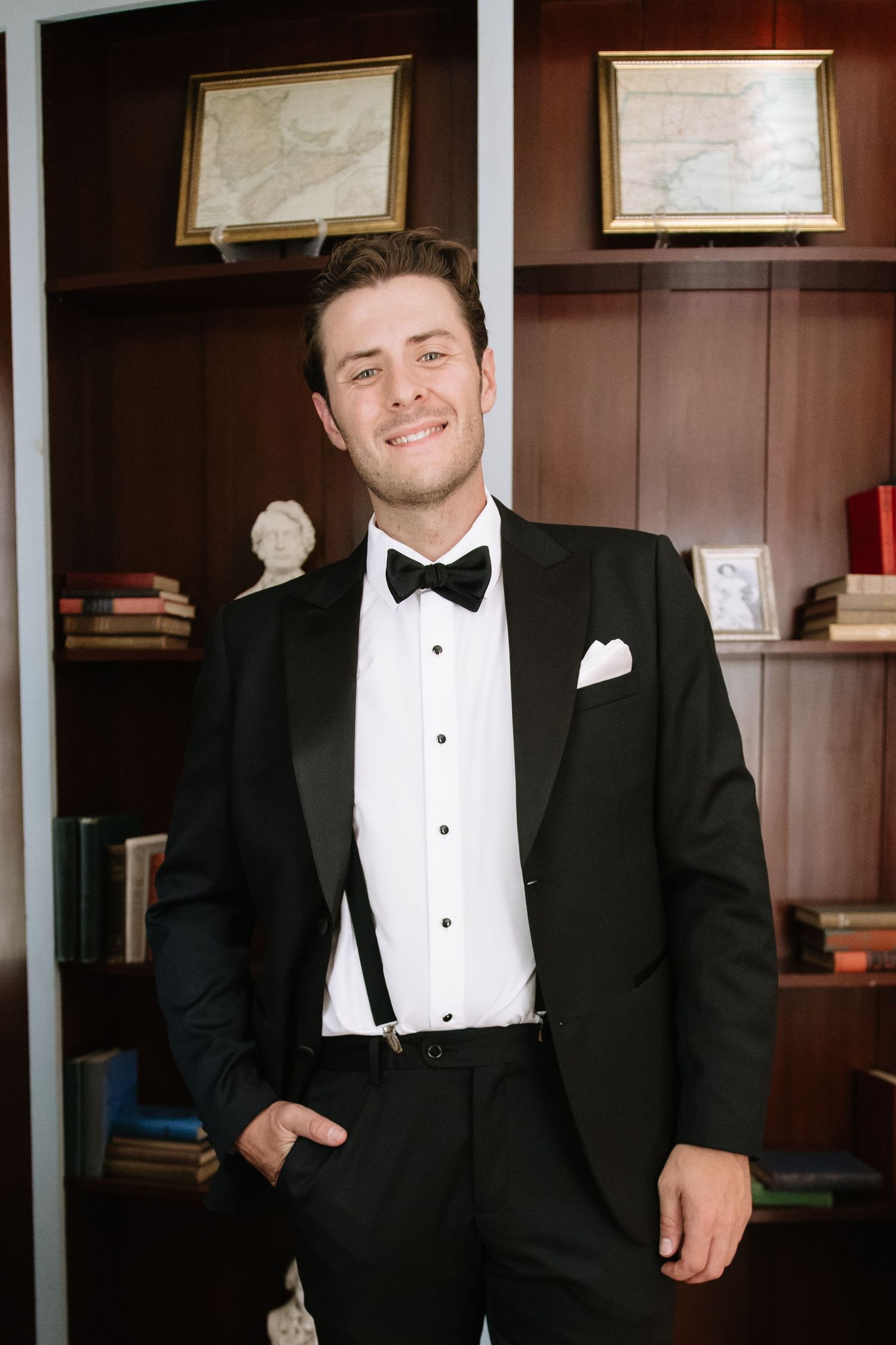 Man in formal attire stands confidently against wood-paneled wall with framed artwork.