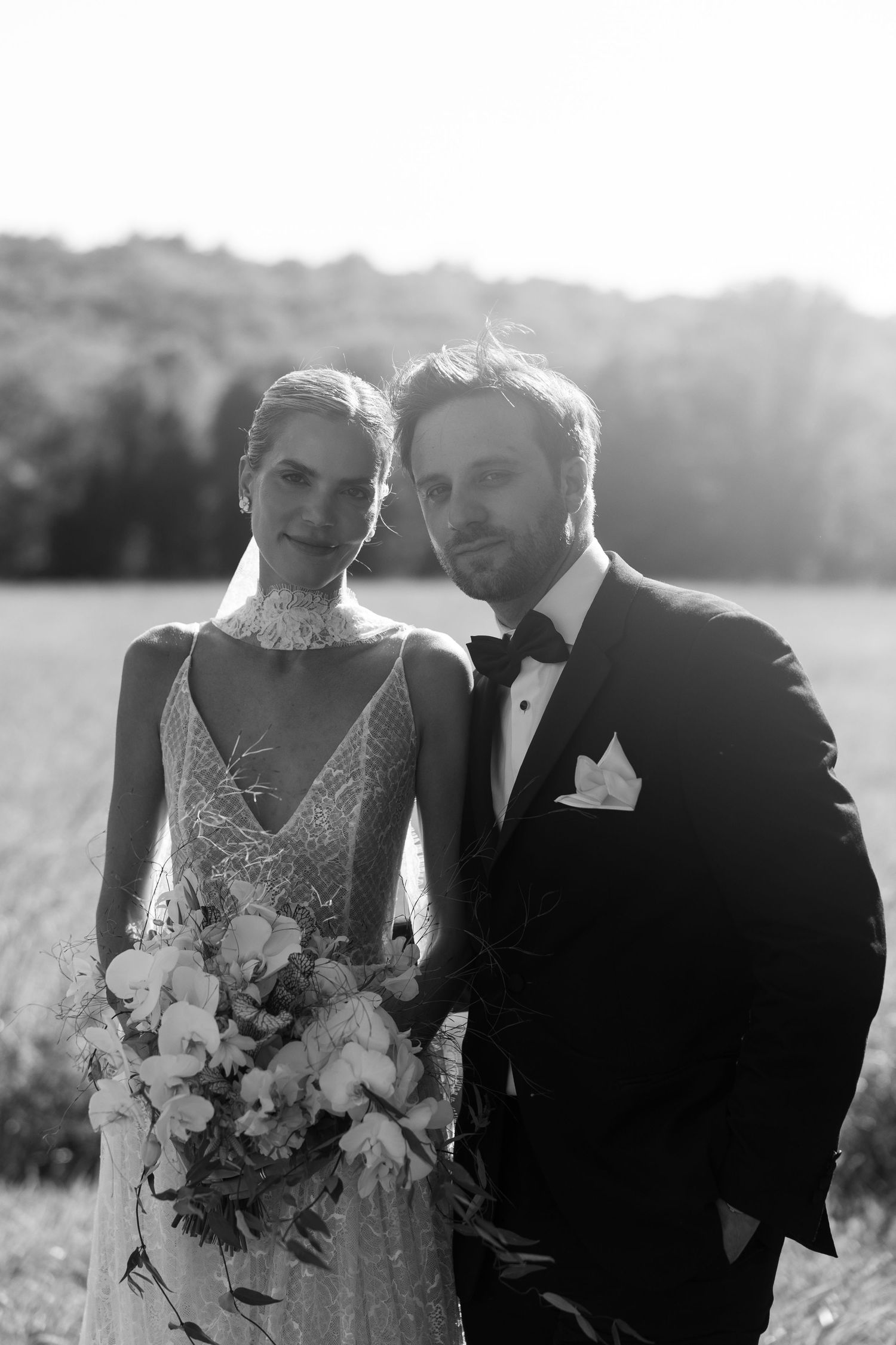 Black and white photo of elegant bride and groom posing in a field, bride holding a lush floral bouquet.