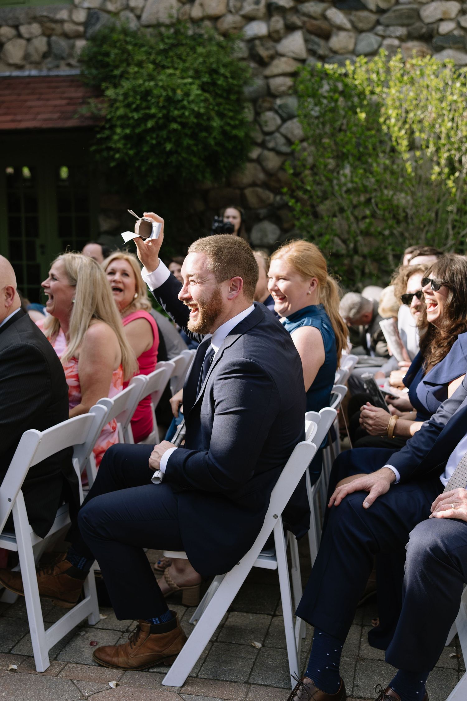 Wedding guests laughing and celebrating during an outdoor ceremony, man in navy suit raising hand joyfully.