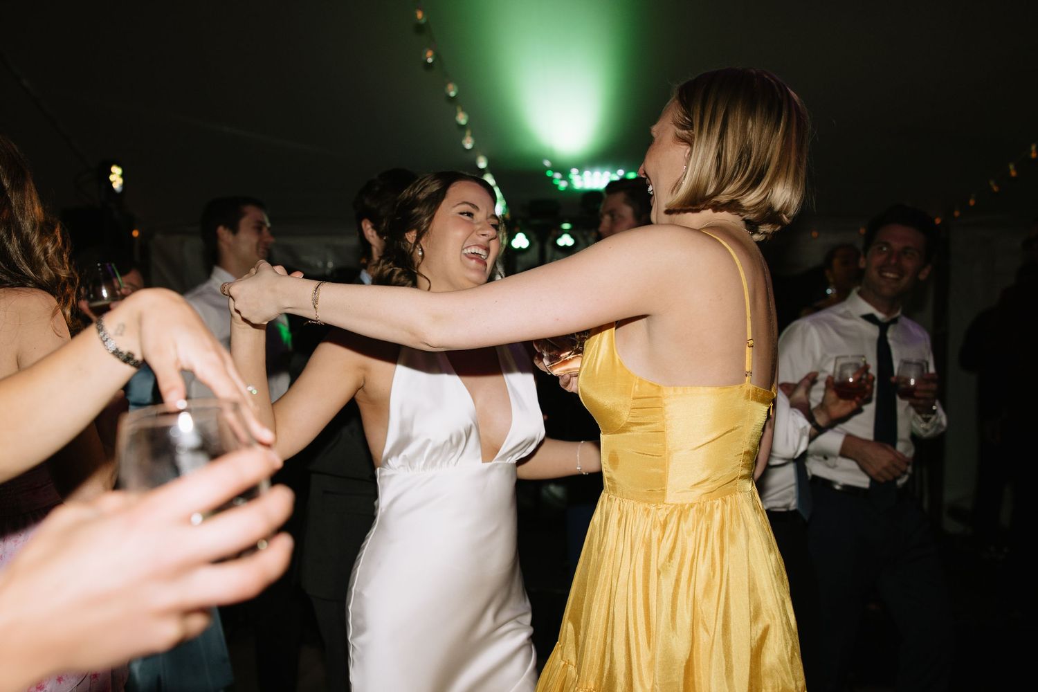 Bride in white gown dancing joyfully with guest in yellow dress at wedding reception.
