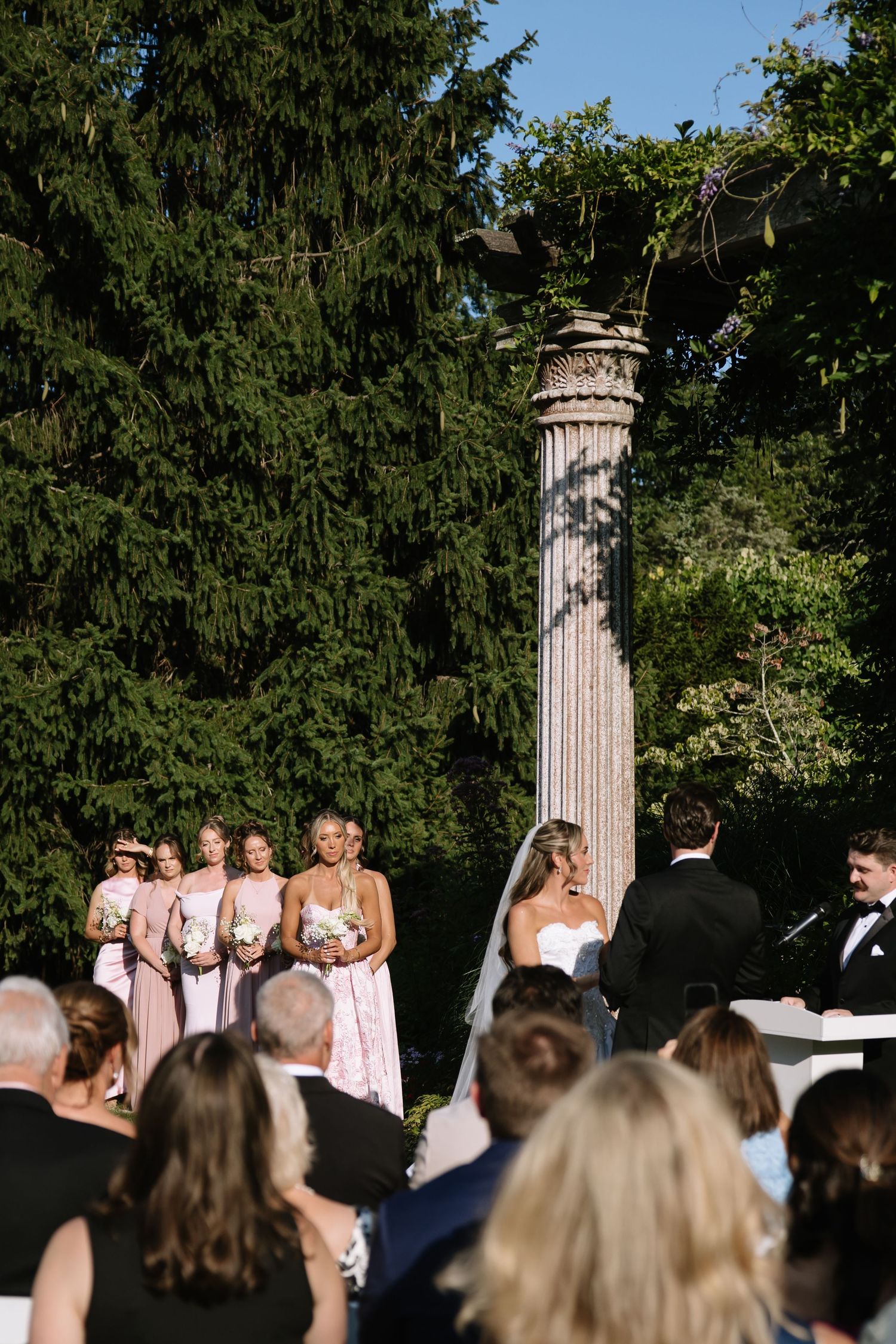 Outdoor wedding ceremony with bridesmaids in pink dresses standing near a stone column surrounded by lush green trees.