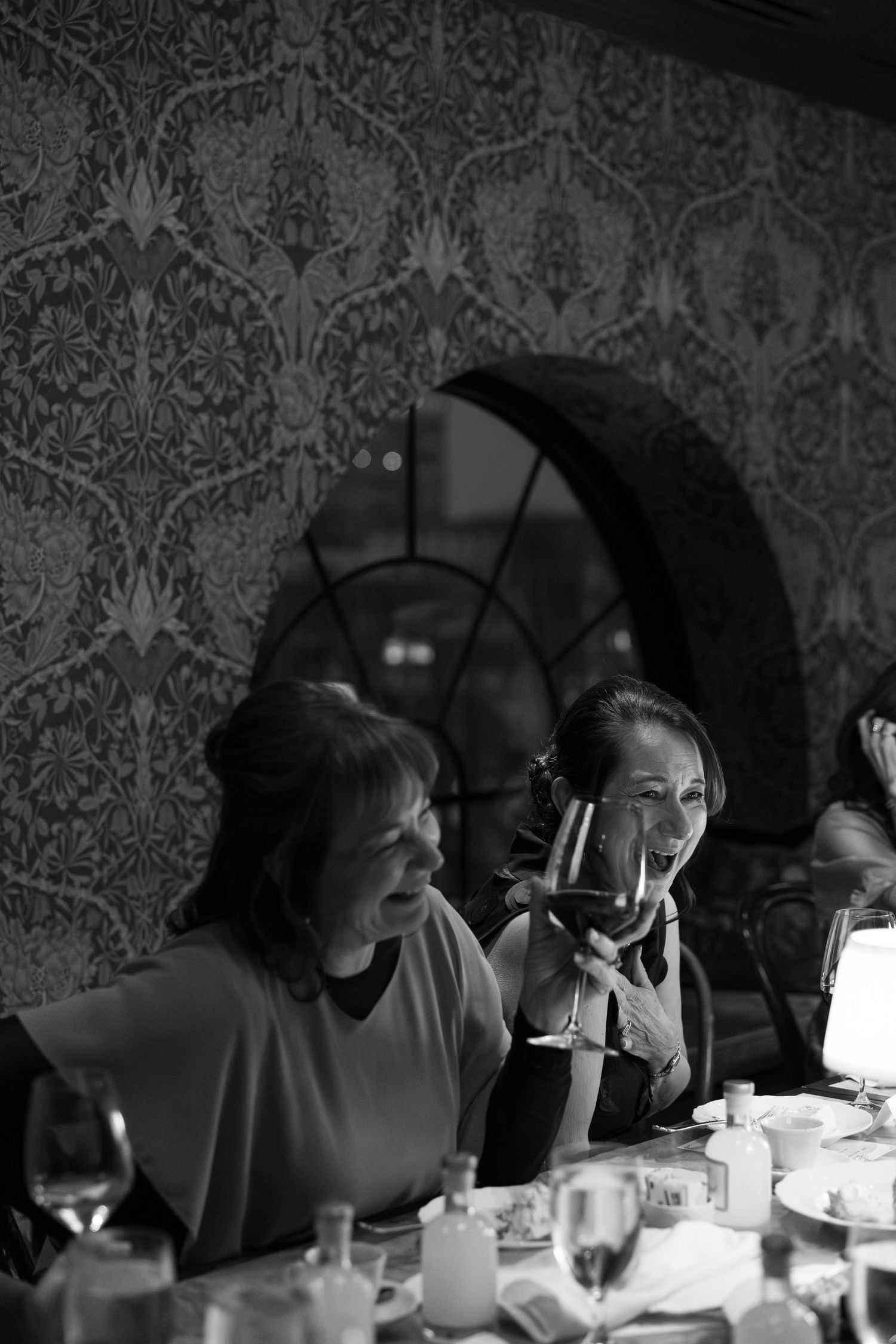 Two women laugh and toast with wine glasses at elegant wedding reception dinner.