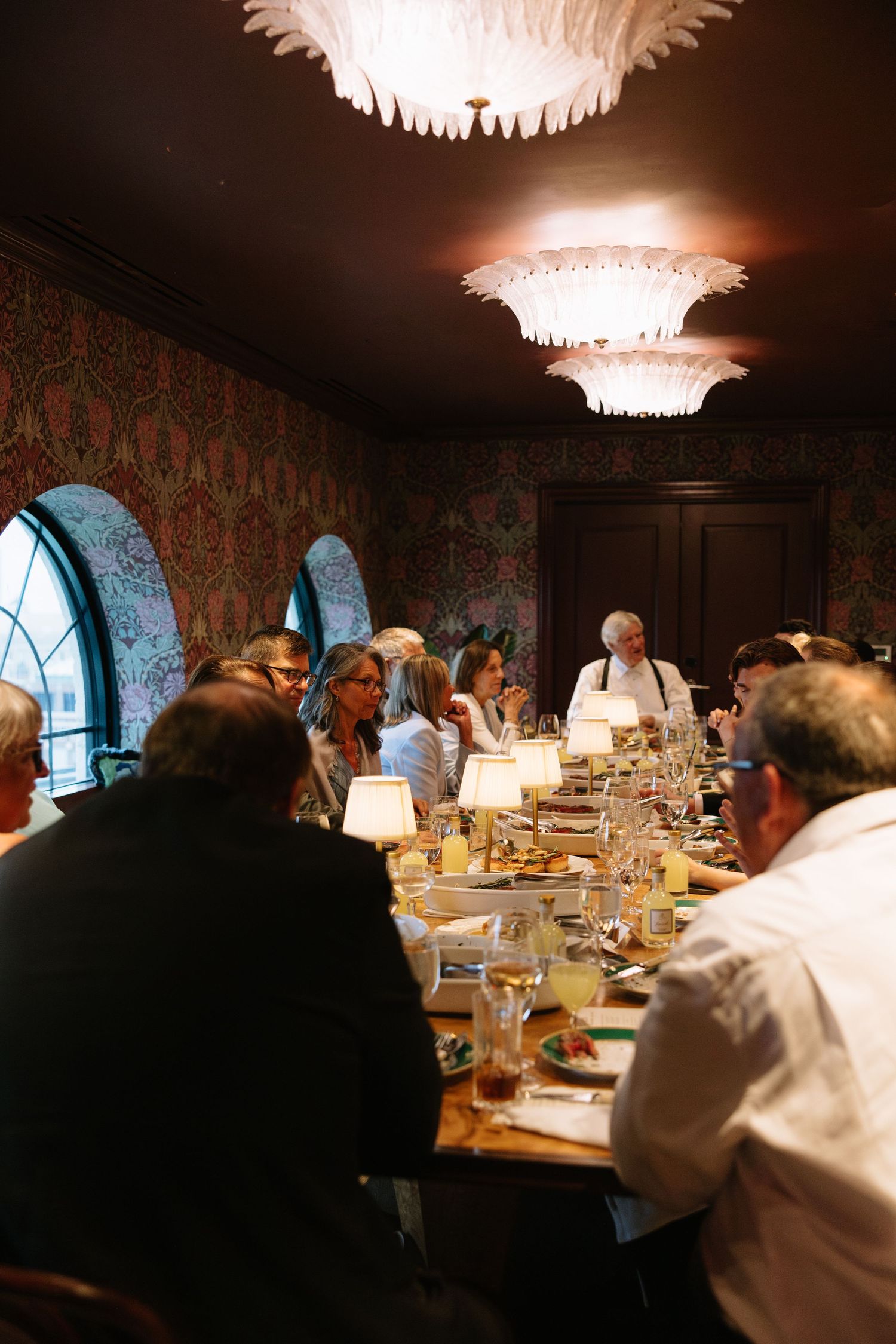 Wedding guests dine at long table in ornate room with floral wallpaper and feathered chandelier overhead.
