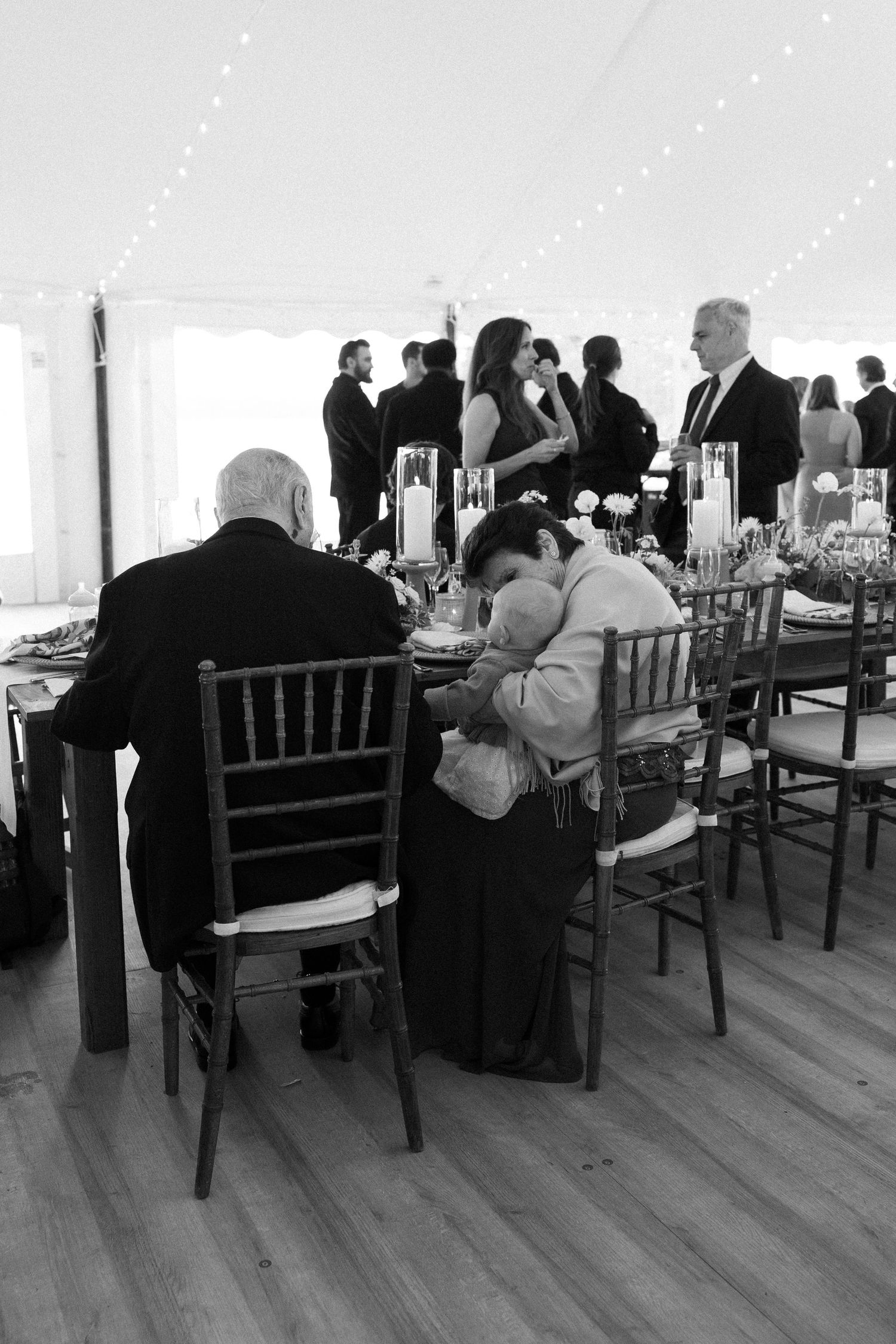 Black and white photo of wedding guests seated at reception tables inside a elegant tent venue with string lights overhead.