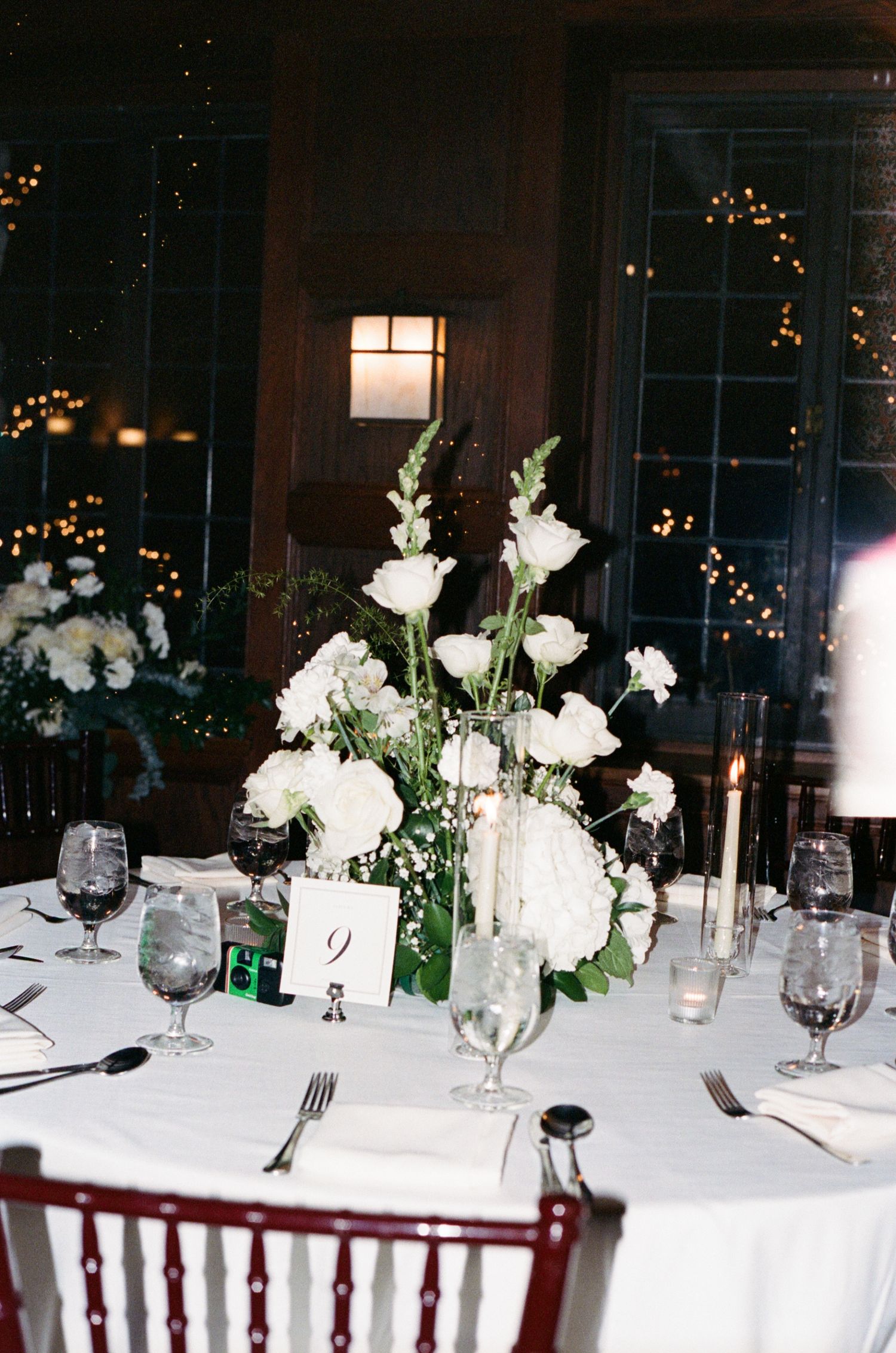 Elegant wedding reception table with white floral centerpiece, candlelight, and dark wood Chiavari chairs in a dimly lit venue.